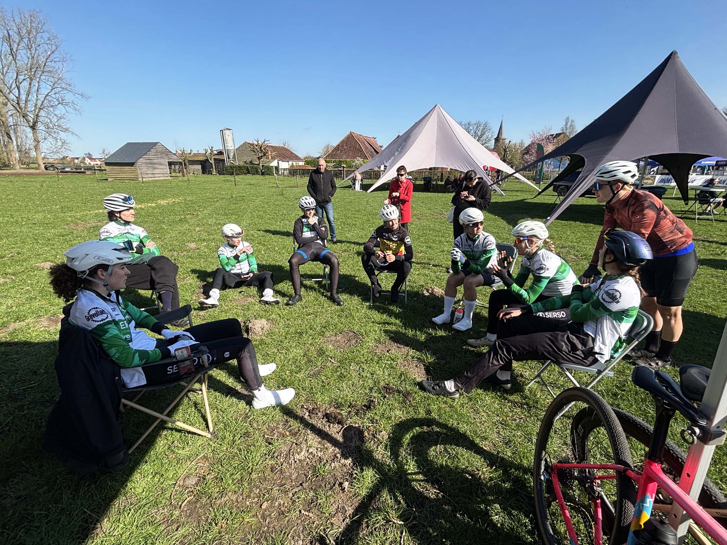 Groep jonge fietsers met helmen zitten op stoelen in een open groene ruimte, er zijn tenten, mensen en huizen op de achtergrond, onder een blauwe hemel.