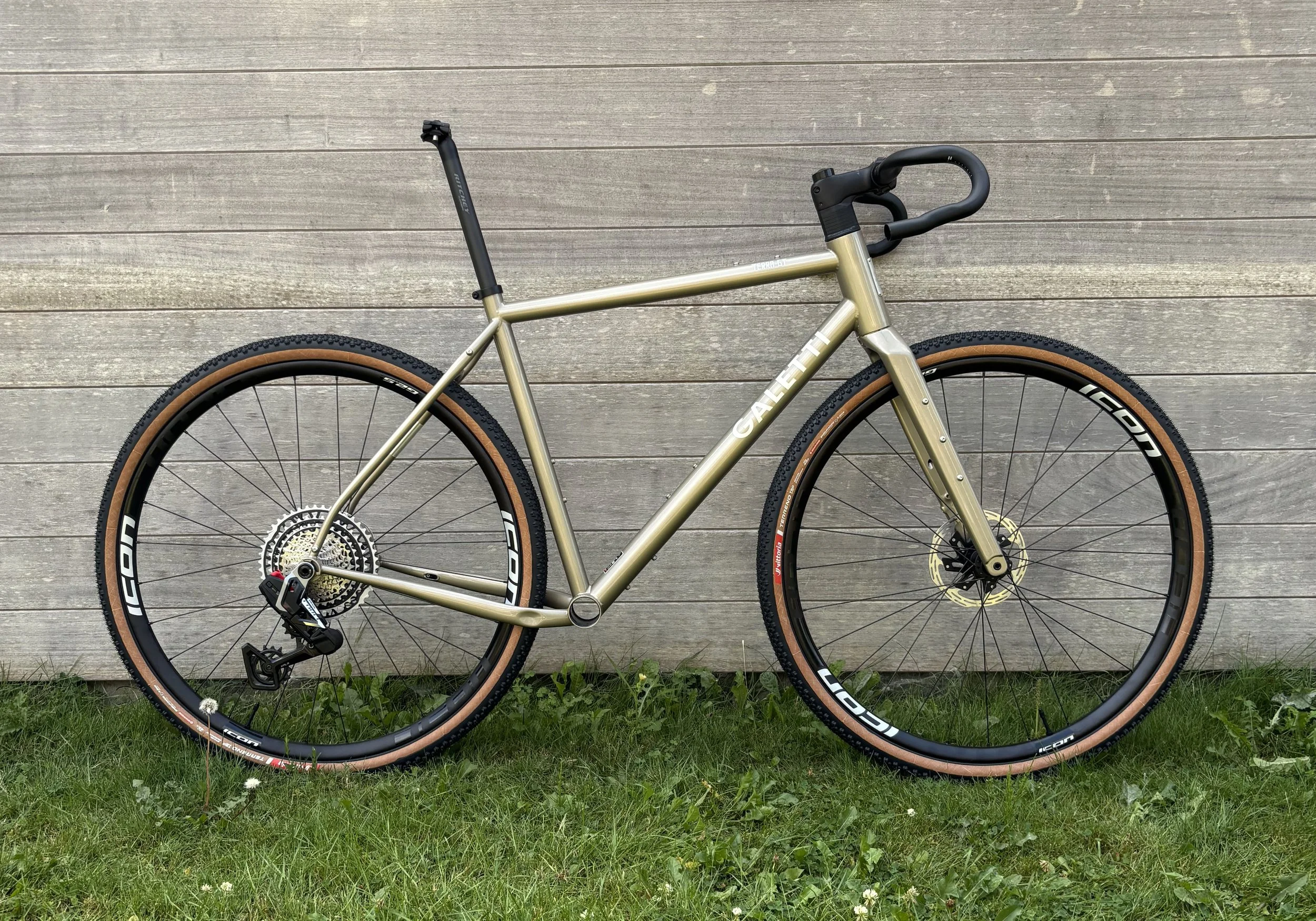 Een gravelbike met een legergroene framekleur, vlakke banden, en zwarte onderdelen, leunend tegen een houten muur met gras op de grond ervoor.