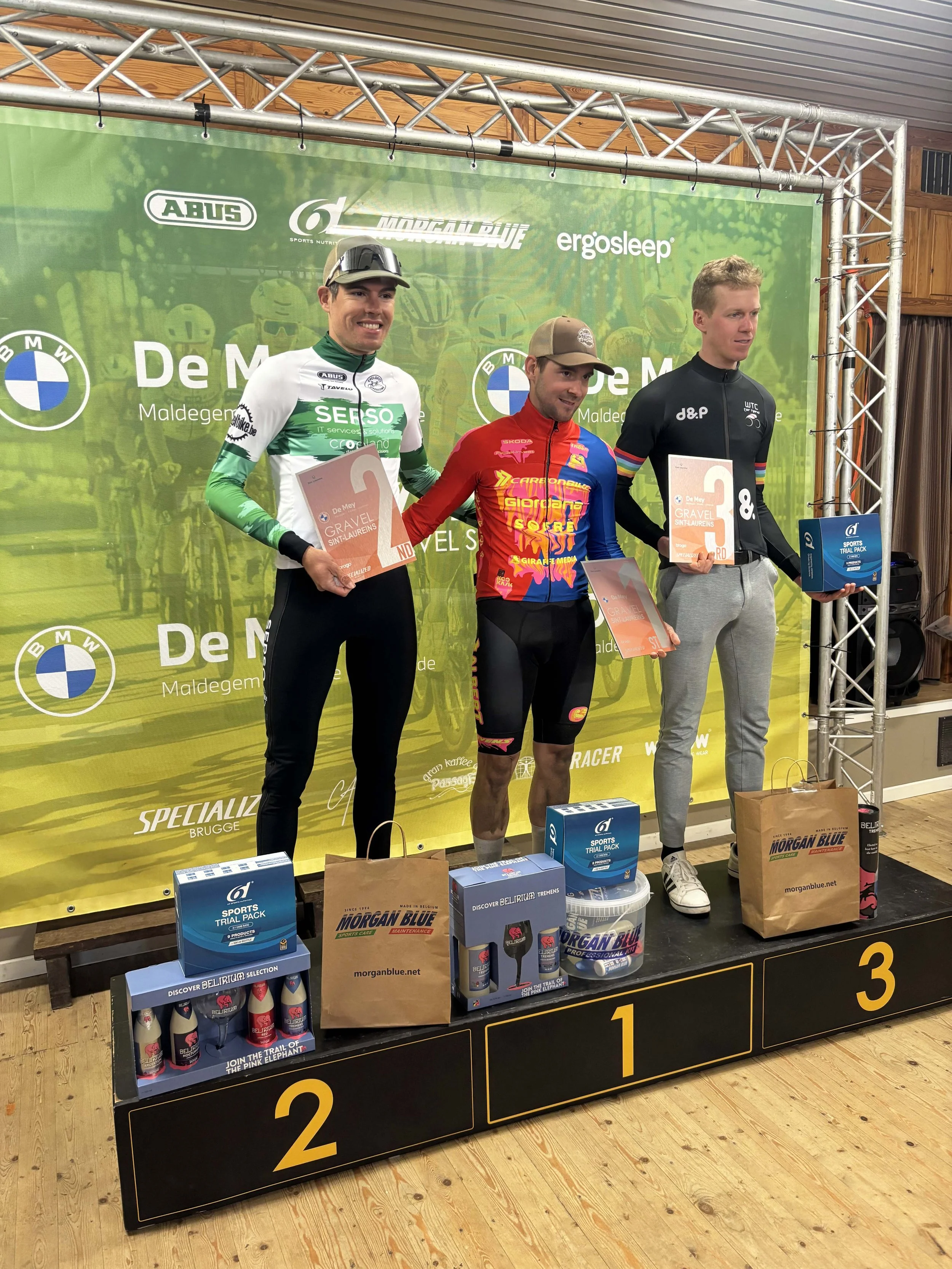 Drie wielrenners staan op het podium tijdens een prijsuitreiking, elk met een certificaat of prijs in de hand. De eerste plaats is bezet door een man in een groene en witte wieleroutfit, de tweede door een man in een rood, blauw en geel wieleroutfit, en de derde door een man in een zwarte wieleroutfit. Op de voorgrond staan dozen en cadeautassen.