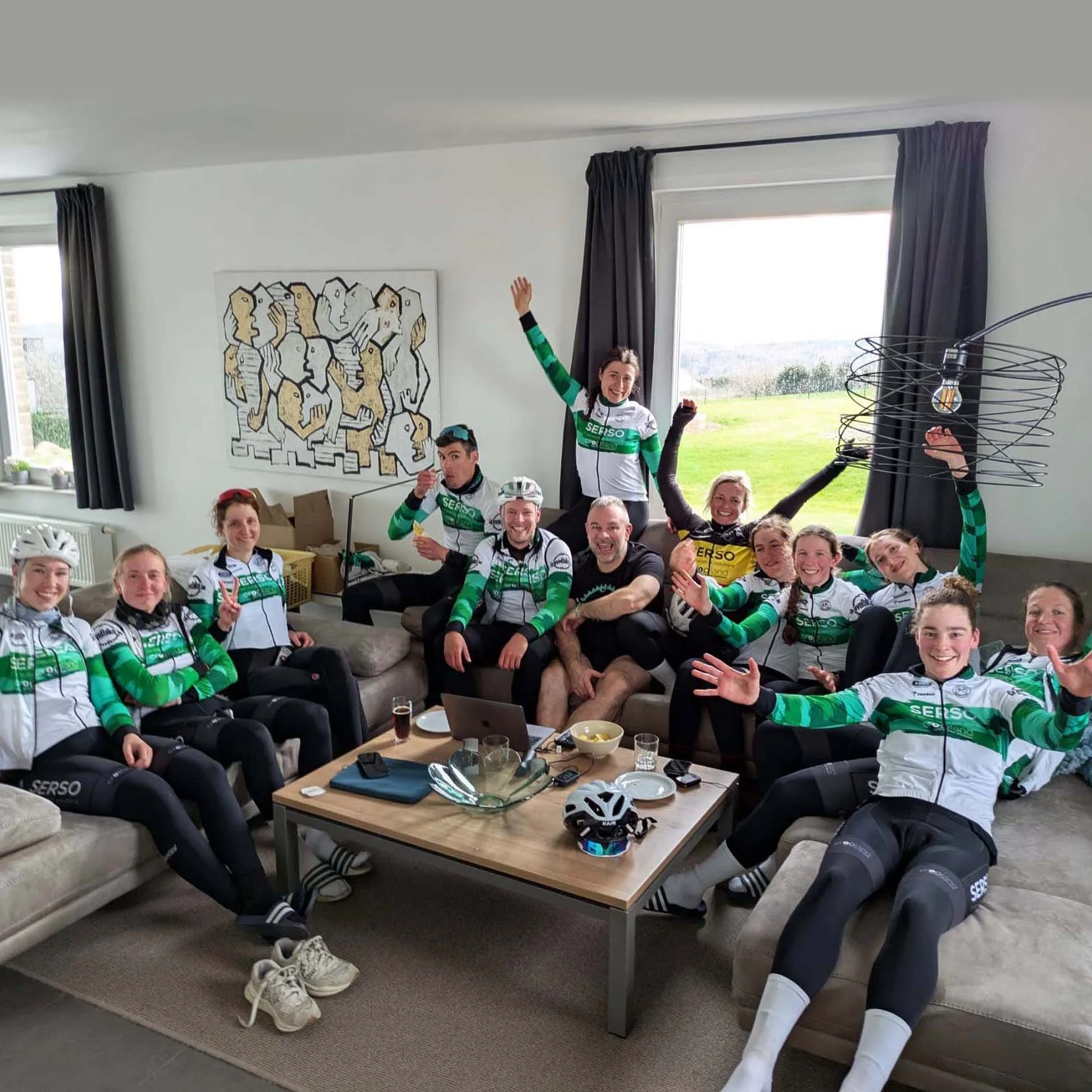 Een groep mensen in wielerkleding zit en hangt ontspannen in een woonkamer met grote ramen, terwijl ze genieten van hapjes en drankjes.