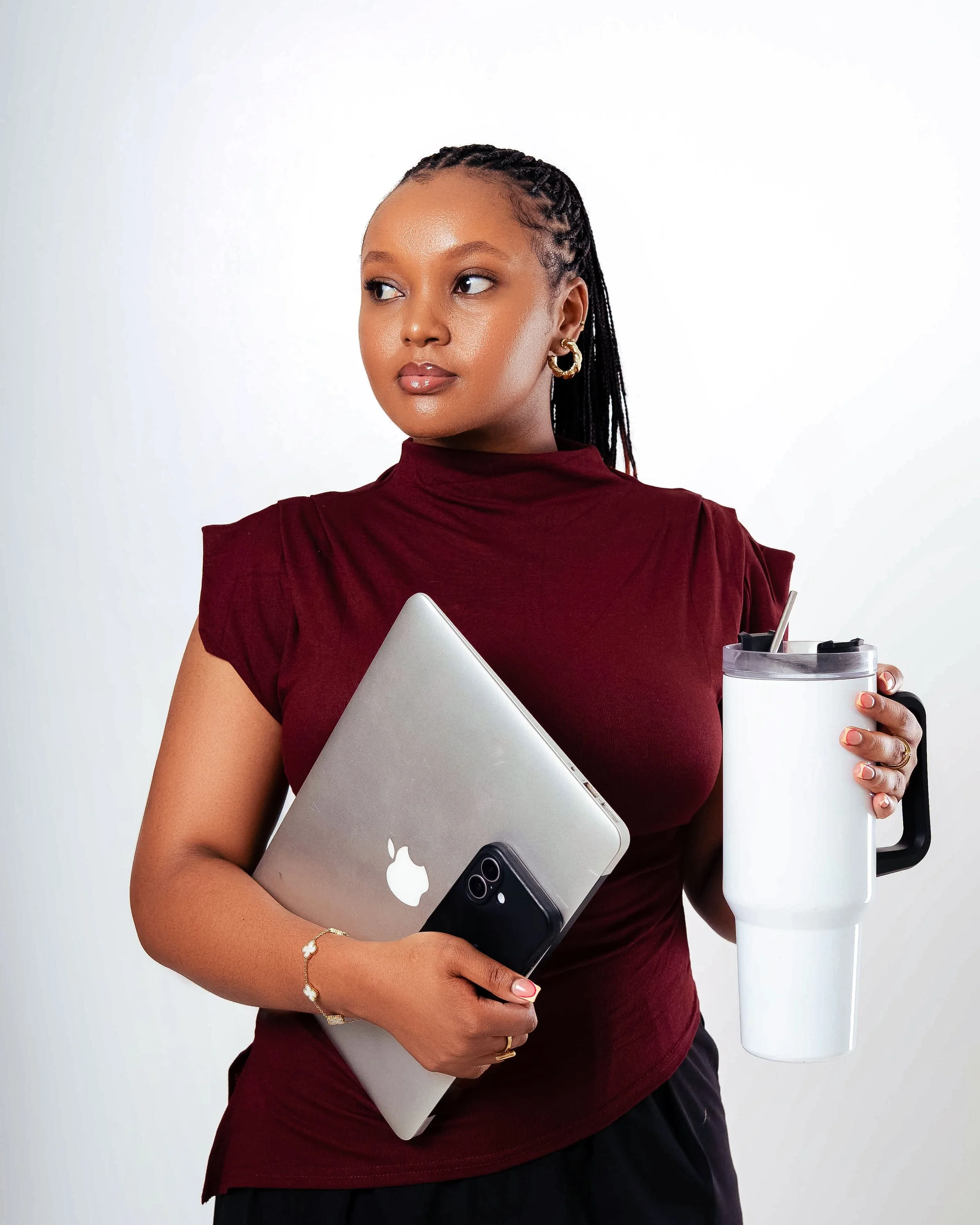 A woman holds a silver MacBook and a black iPhone in her arms, with a laptop, a white tumbler, and a pen in her hand, standing against a plain white background.