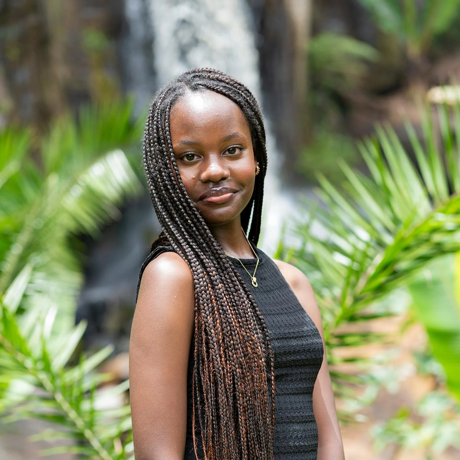 A young woman with long braided hair standing outdoors in front of a waterfall and lush greenery, wearing a black sleeveless dress, gold jewelry, and smiling gently.