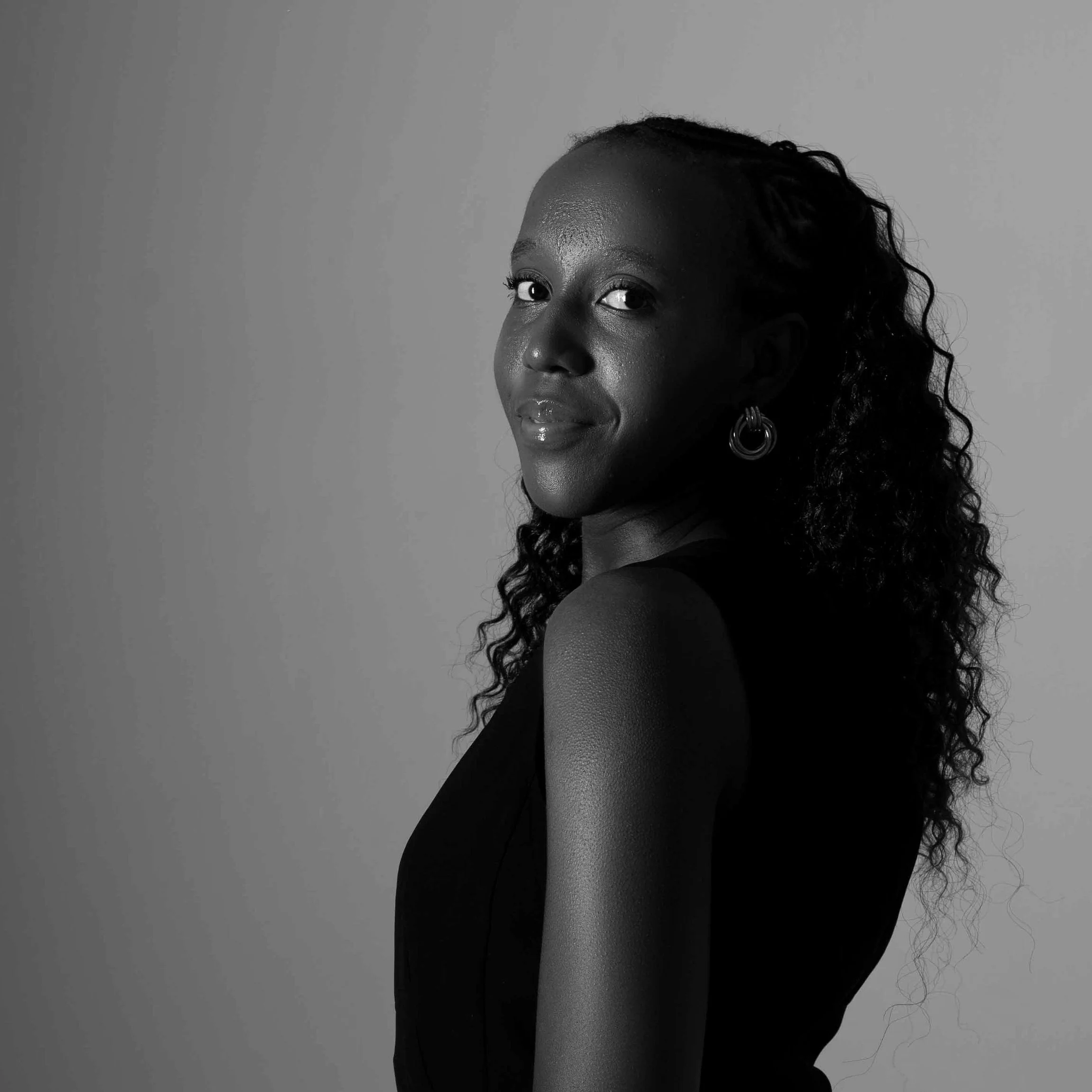 A black and white photo of a woman with dark skin, curly hair, and earrings, looking over her shoulder against a plain background.