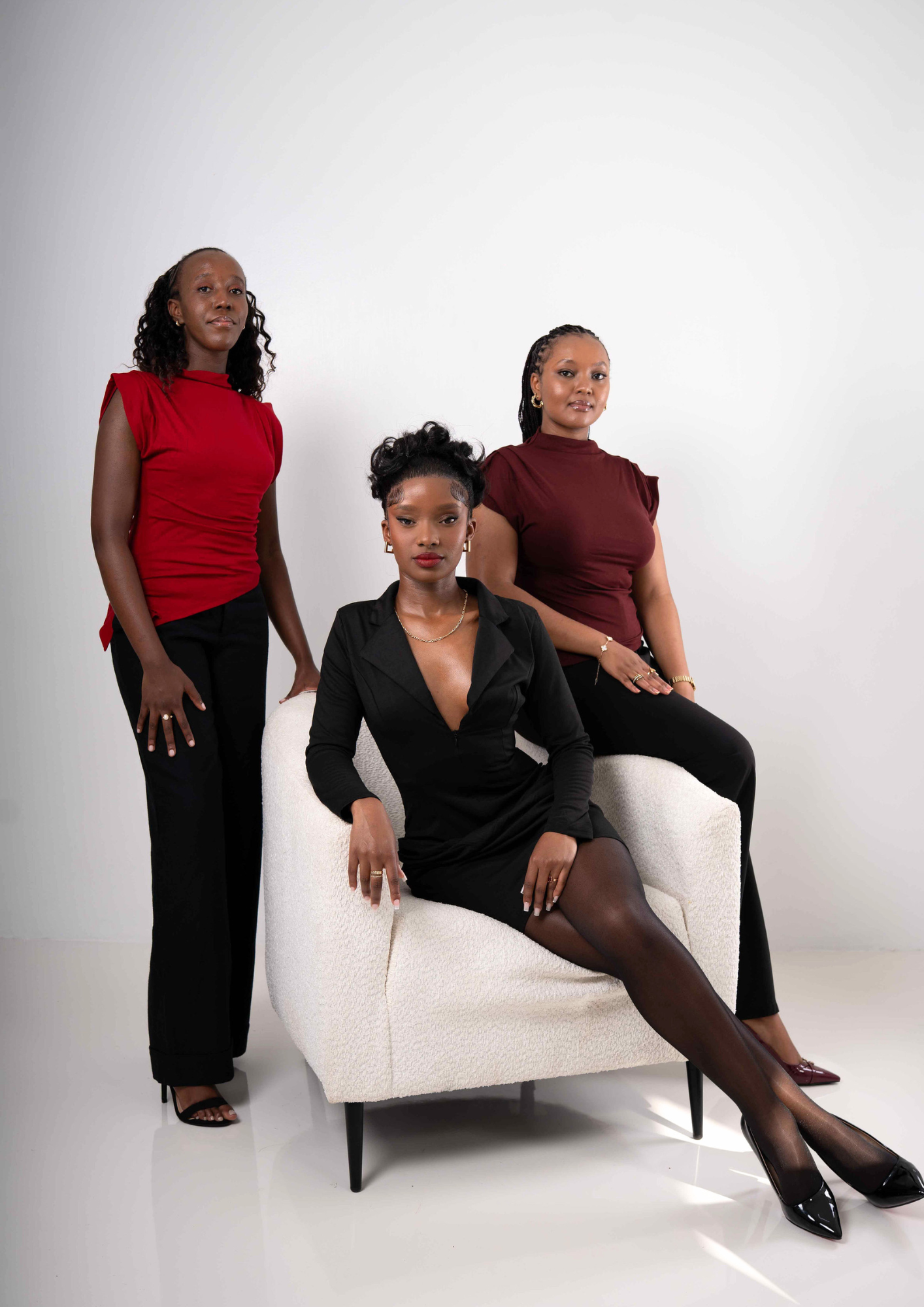Four women with dark skin and dressed stylishly pose in a studio with a plain white background. One woman is seated on a white textured armchair with her legs crossed, while the other three stand beside her, two on her right and one on her left.