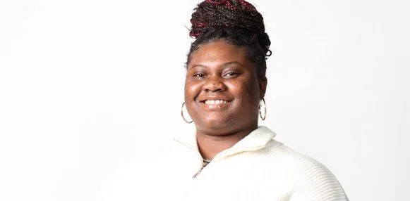 A woman with dark hair styled in braids, smiling, wearing a cream-colored top against a white background.