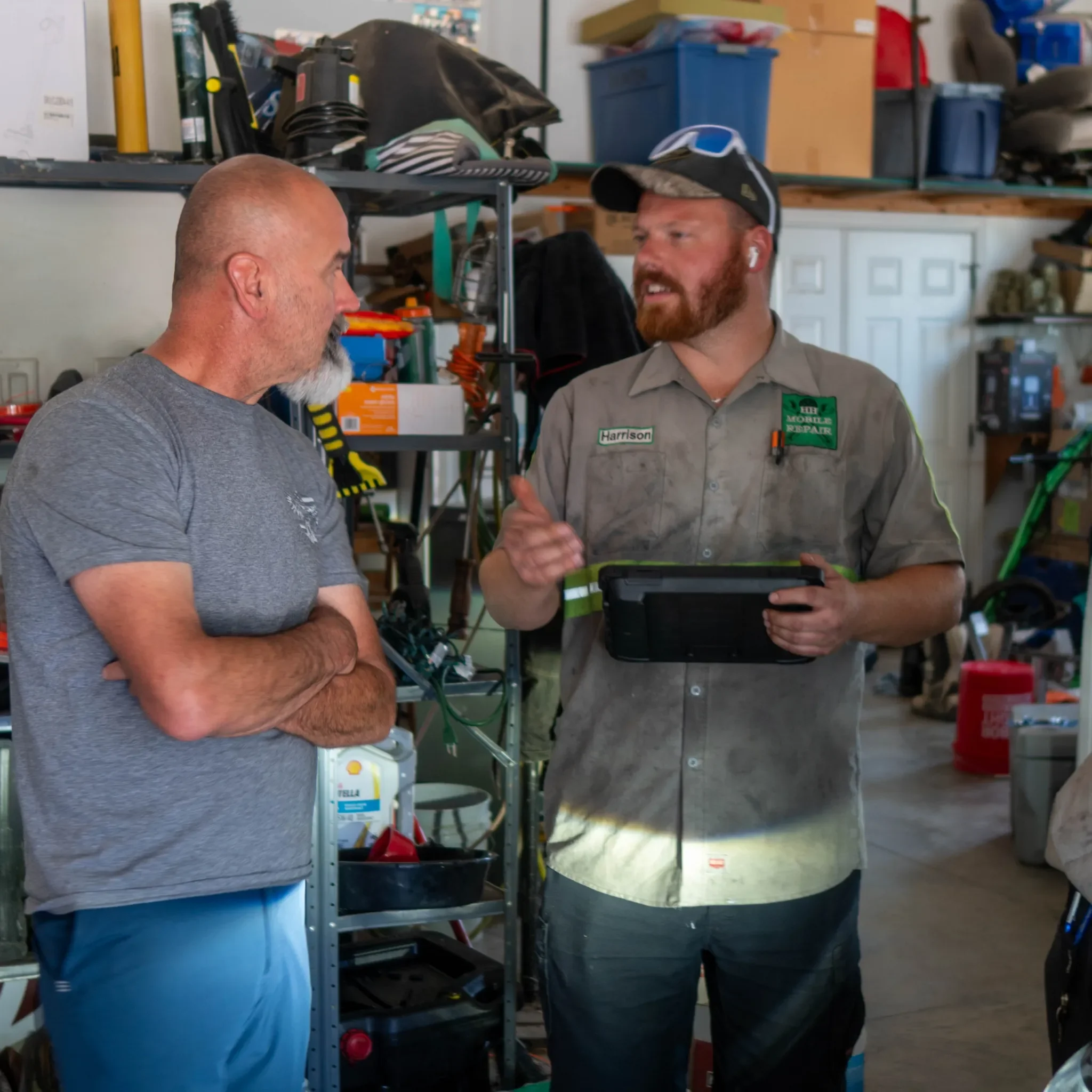 Two men talking in a garage or workshop, one with a bald head and gray beard, wearing a gray shirt; the other with a red beard, wearing a gray uniform with a name tag and a cap and sunglasses on his head, holding a tablet or phone.