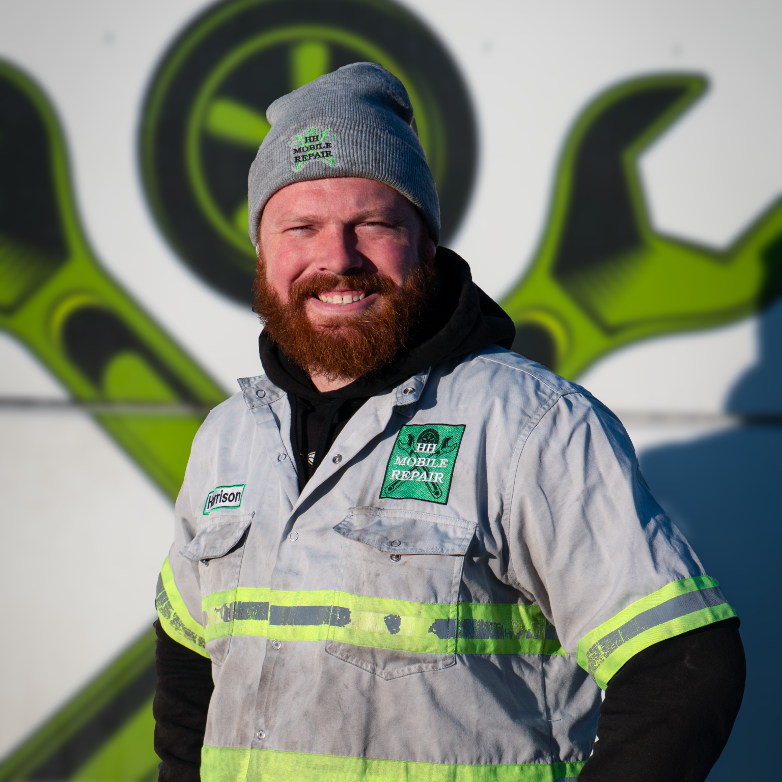 A smiling man with a beard wearing a gray beanie, a light gray work jacket with a 'Mobile Repair' patch, and a black hoodie underneath. He is standing outdoors in front of a large sign featuring a green and black wrench and gear logo.