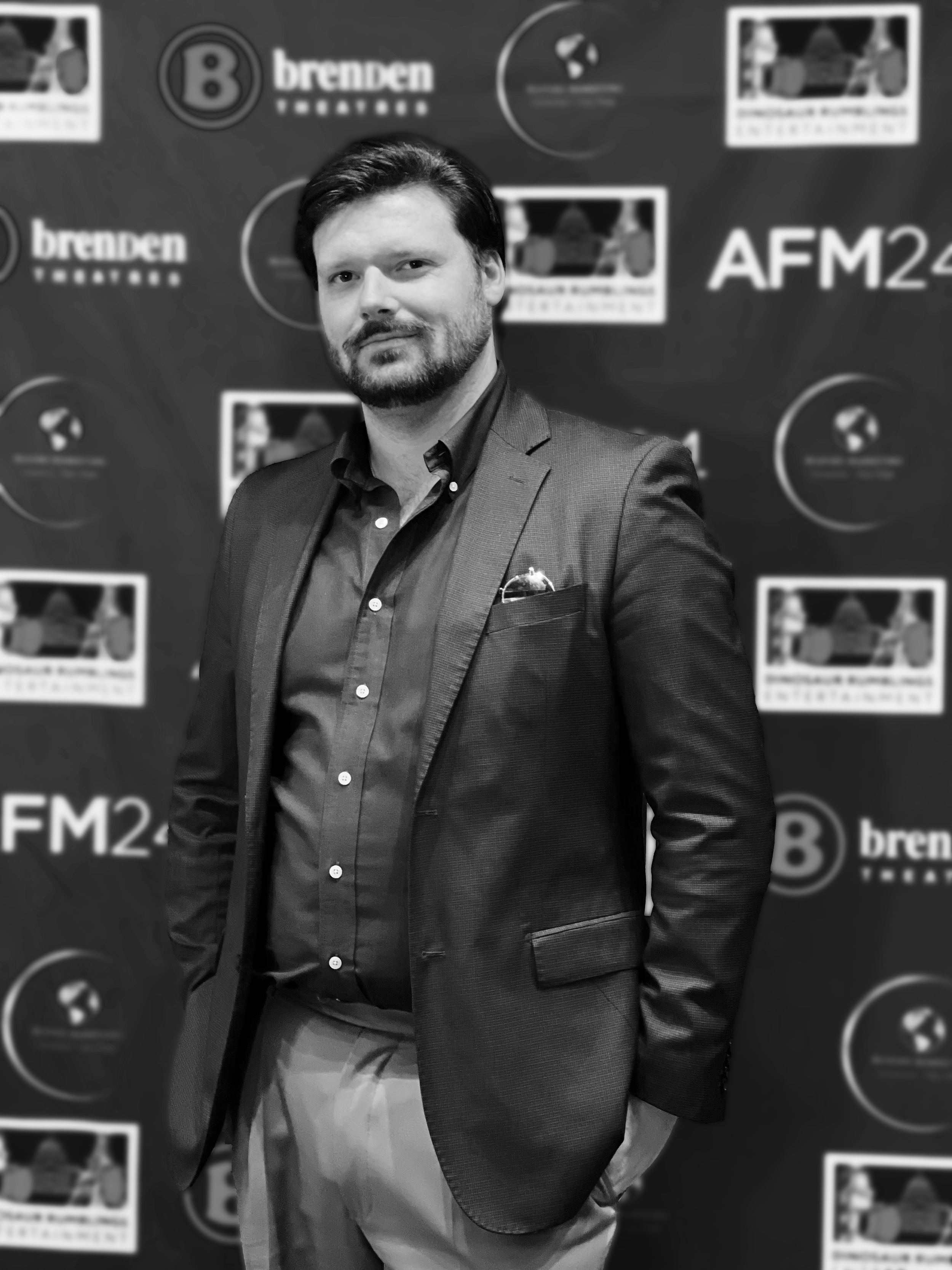 A man in a suit jacket and dress shirt standing in front of a backdrop with logos and text, including 'brenden TELEVISION,' 'AFM24,' and 'B' in circles.