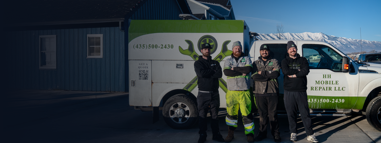 Four male repair technicians stand in front of a white and green mobile repair truck outside a building with mountains in the background, with one wearing a high-visibility uniform.