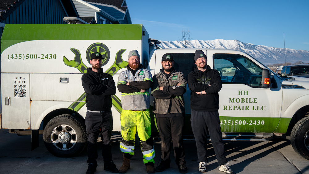 Four men standing in front of a white and green mobile repair truck with mountain range in the background. The truck has a logo with tools and a QR code, and the text "HH Mobile Repair LLC" and a phone number. One man is in a high-visibility yellow work suit, and the others are in dark clothing and hats.