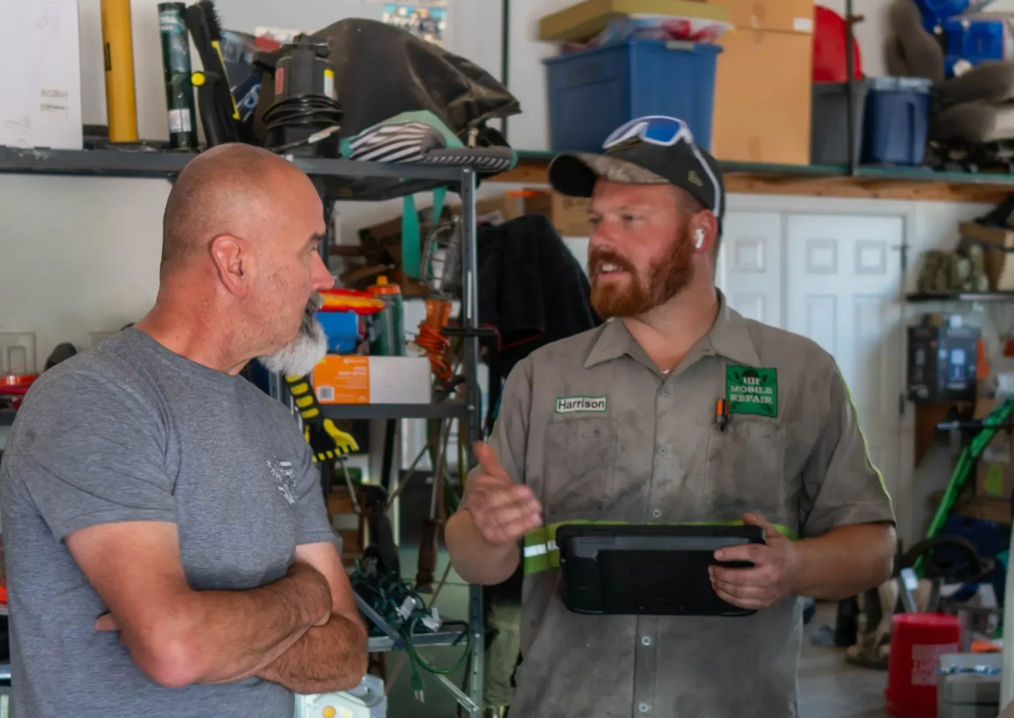 Two men are engaged in conversation inside a garage or workshop. One man has a shaved head and gray t-shirt, the other has a beard, wears a work uniform with a name tag 'Harrison' and appears to be explaining something using a tablet. Shelves with boxes and tools are visible in the background.