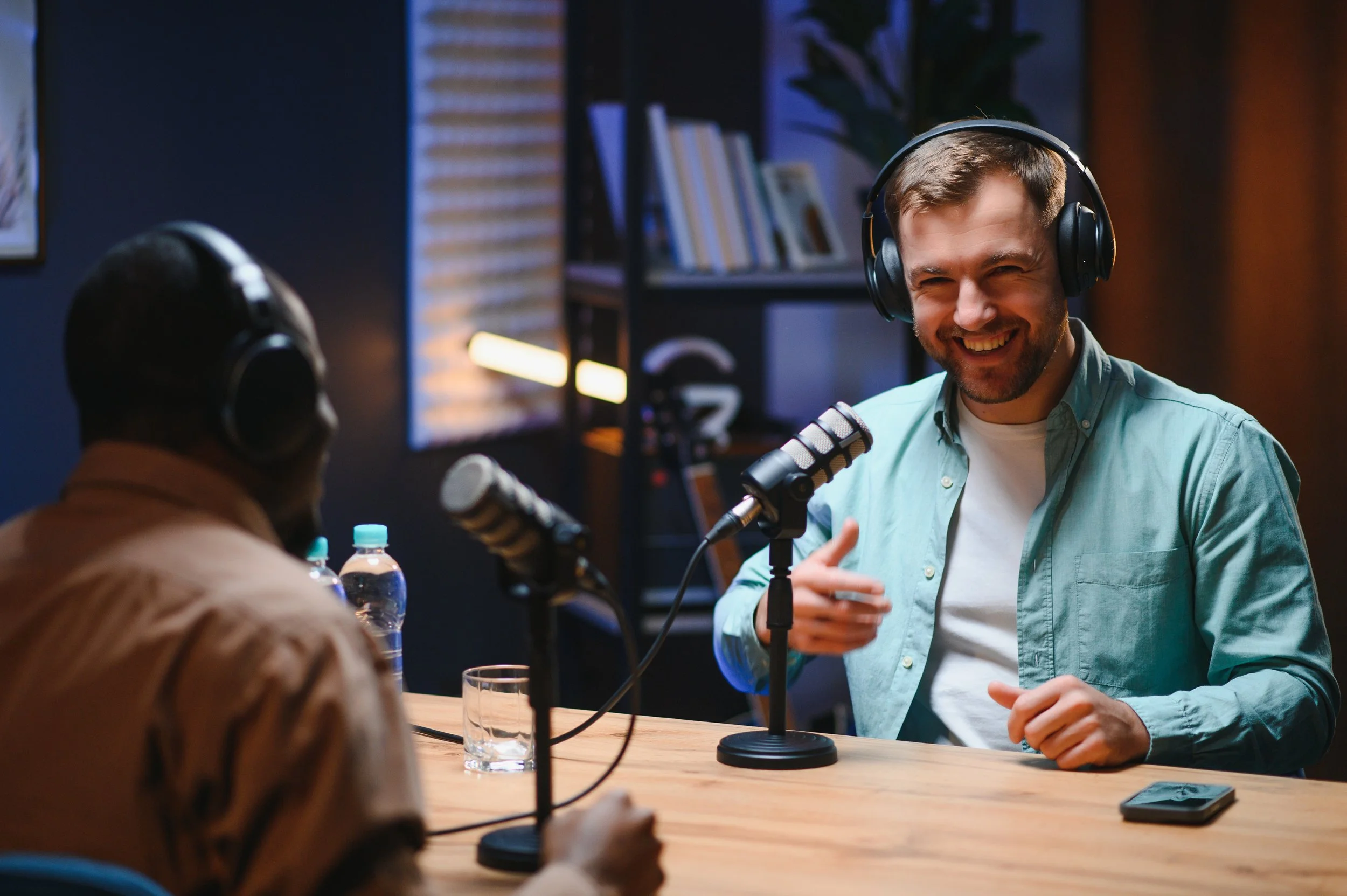 A man with a beard and wearing headphones is smiling and speaking into a microphone during a radio or podcast interview in a studio. There are people with headphones sitting at a table in front of him, with water bottles and glasses on the table.
