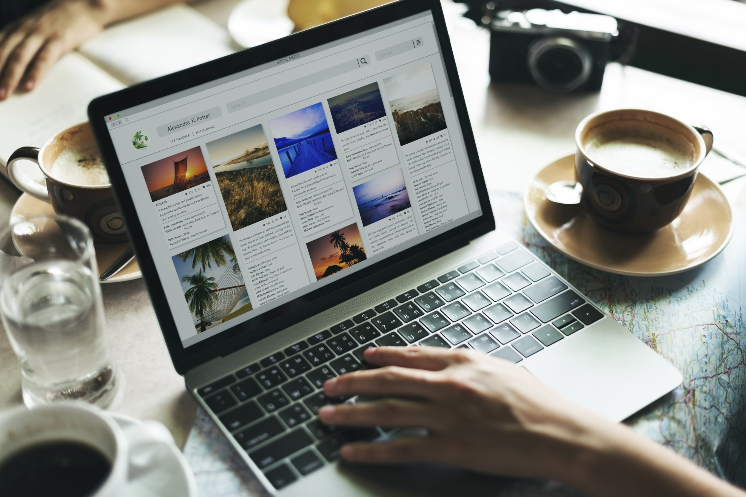 A person viewing a social media page on a laptop with cups of coffee and a camera on the table.