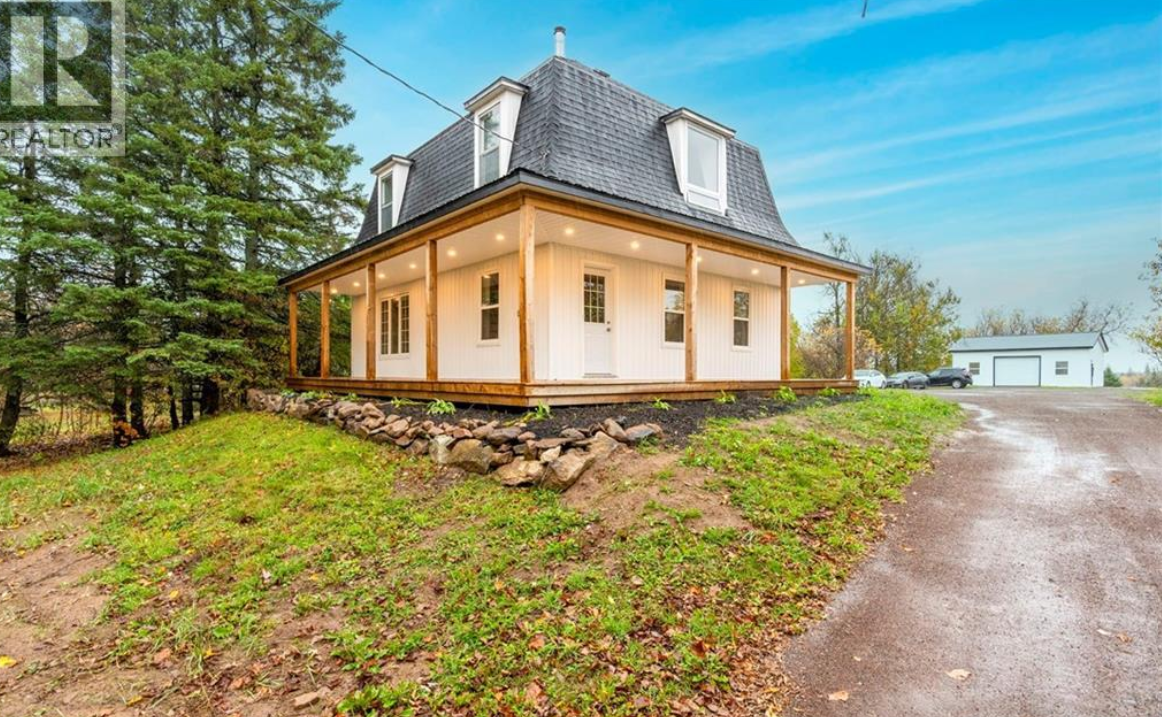 A two-story house with a modern roof, surrounded by trees, with a large porch that has wooden railings and support beams, on a grassy lot with a gravel driveway in the foreground.