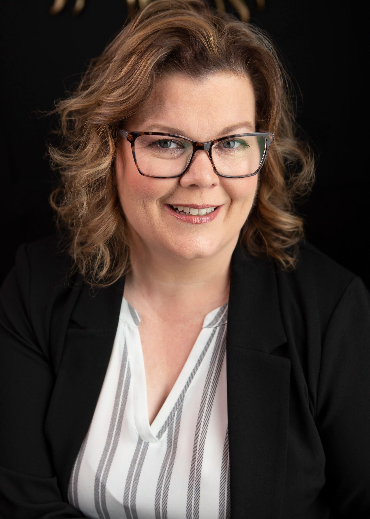 Headshot of a woman with short, curly brown hair, wearing glasses, a black blazer, and a white striped blouse, smiling against a black background.