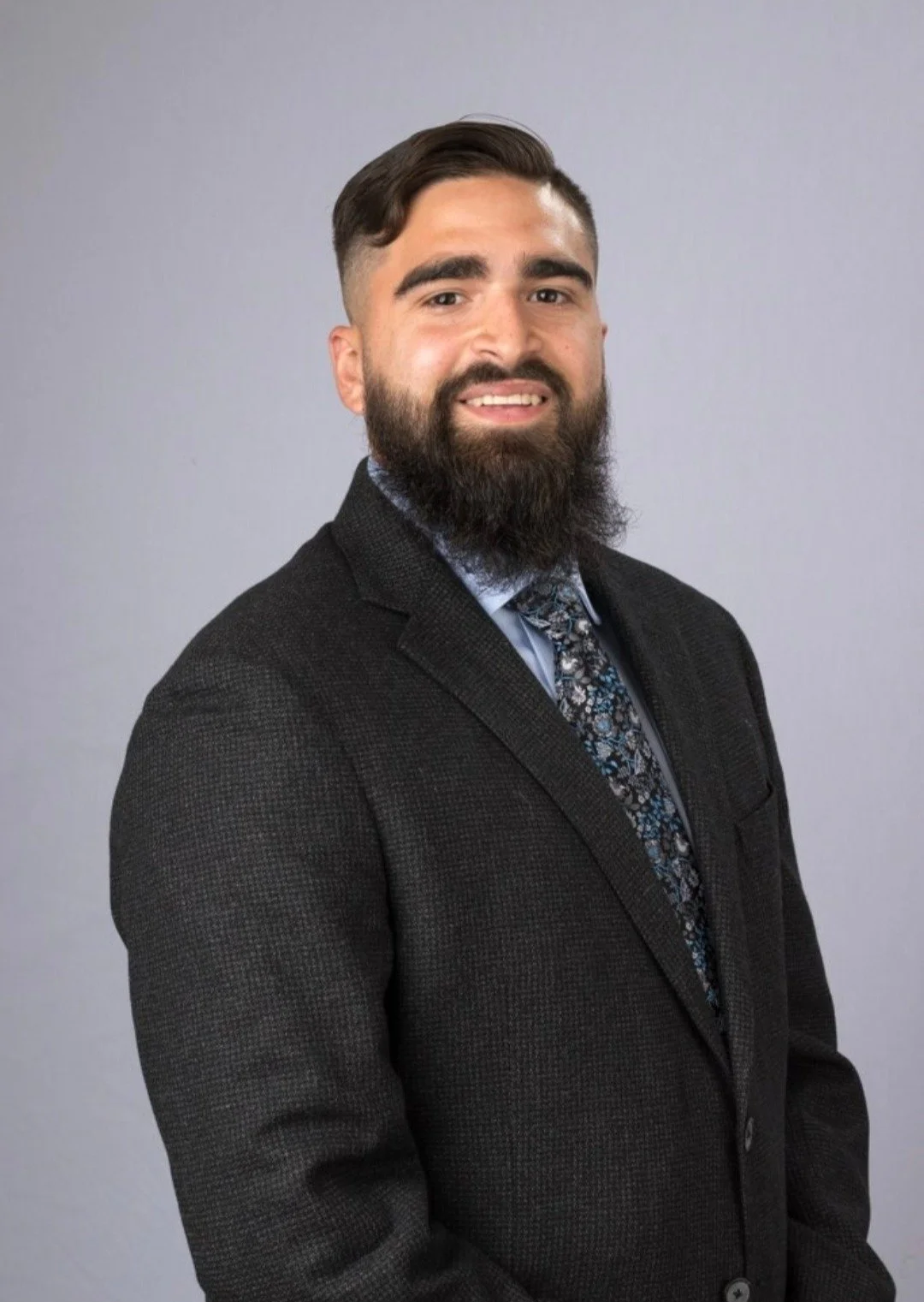 A man with a beard and mustache, wearing a dark blazer, light blue shirt, and a patterned tie, standing against a plain gray background.