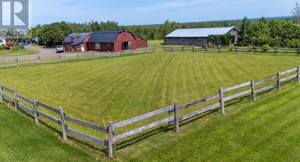 A large grassy lawn enclosed by a wooden fence on a rural farm, with two barns and some trees in the background.