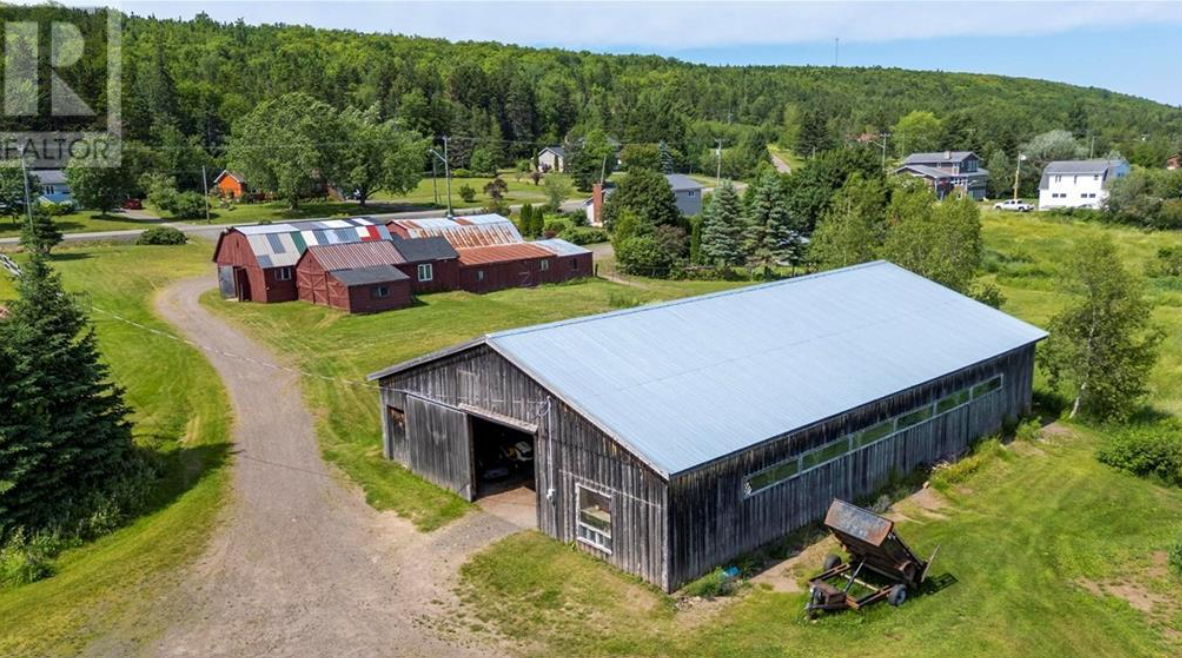 Aerial view of a rural area with two large barns, green grassy fields, trees, and a few houses in the background.