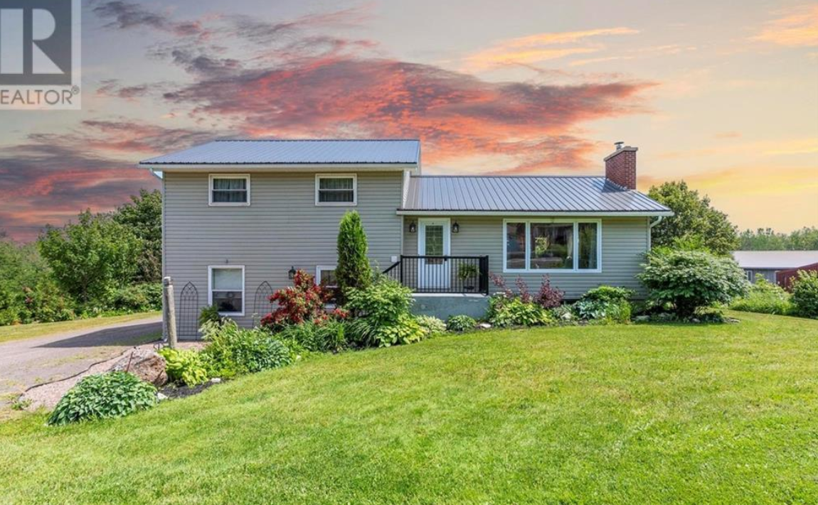 A house with a green lawn and garden, sunset sky with orange clouds, vinyl siding, metal roof, chimney, and a small porch with black railing.