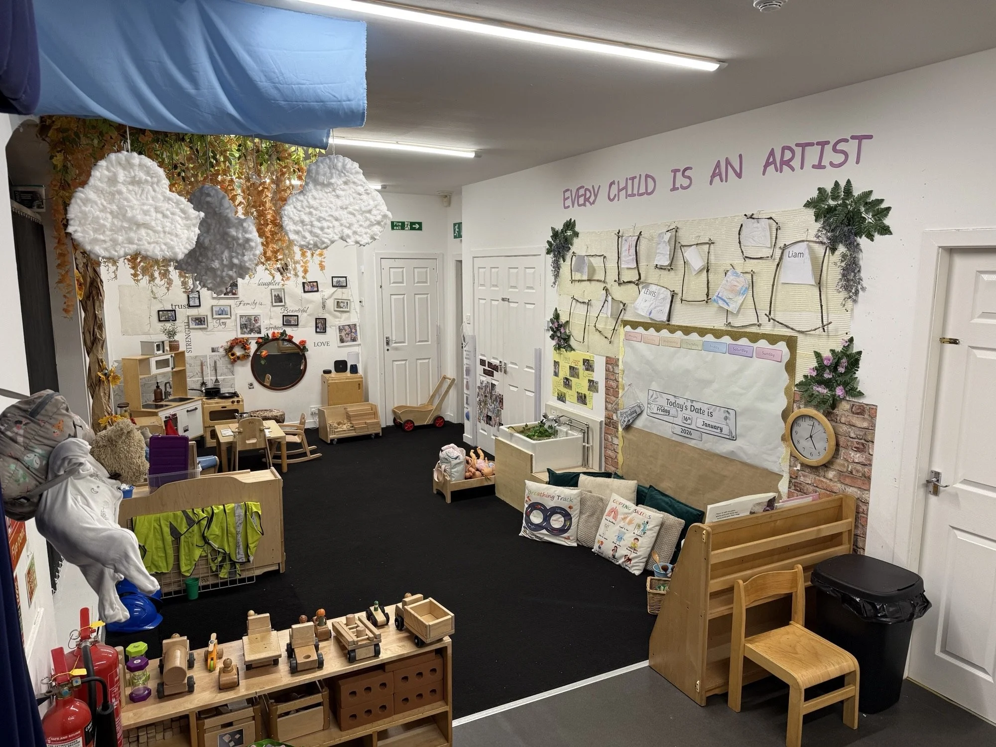 A colorful classroom with hanging cloud decorations, a reading and activity area with cushions, a toy kitchen, and various educational materials and artworks on the walls, including a sign that says "Every Child is an Artist."