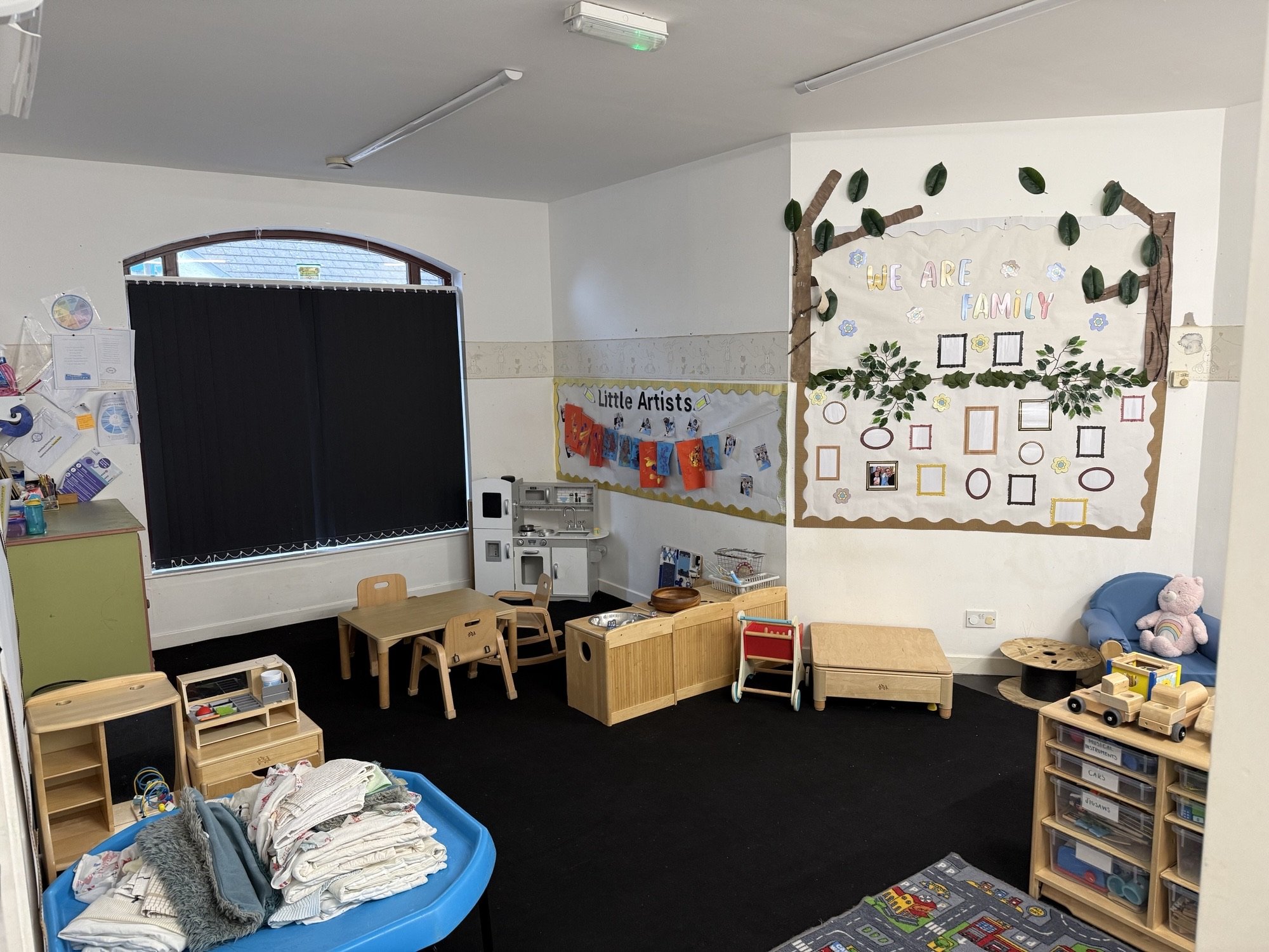 Childcare or preschool classroom with small tables, chairs, toys, and bulletin boards, including a display titled 'We Are Family' and 'Little Artists'.