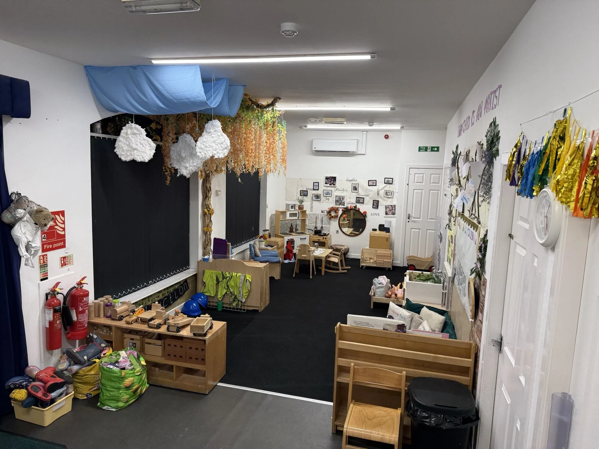 A classroom decorated with autumn-themed and colorful decorations. There are toys and supplies on shelves, with black chalkboards on the left, a small seating area with chairs and cushions, and various wall displays and artwork.