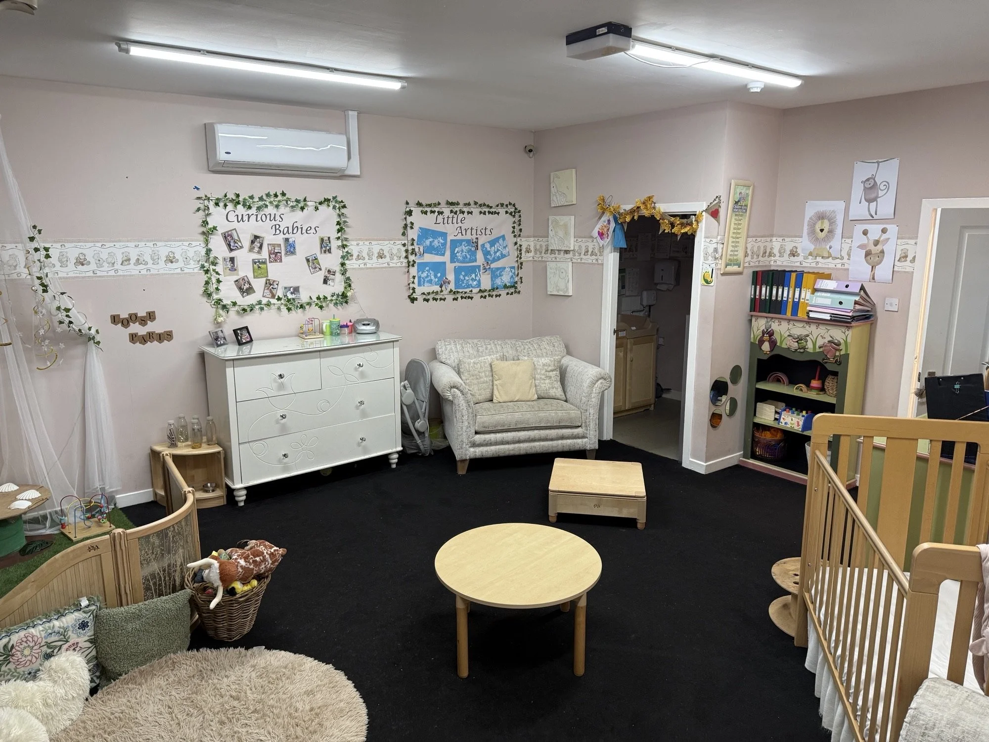 A cozy, well-decorated children's waiting room with a black carpet, small wooden tables, a cream-colored couch, a white dresser, and various children's artwork and decorations on the walls.