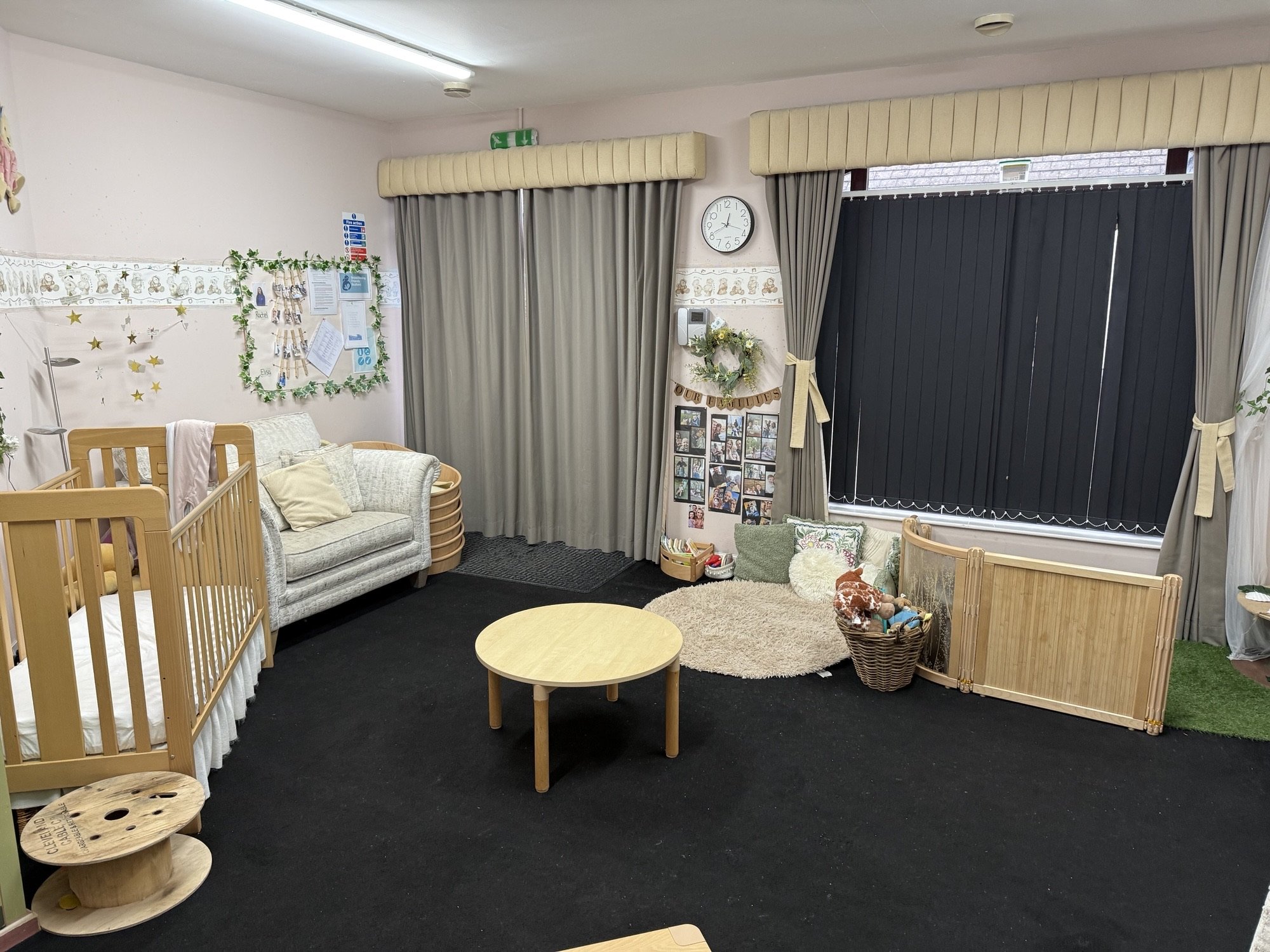 Interior view of a children's playroom with a small couch, a crib, a round table, a basket of toys, a soft rug, and decorated walls with photos and a clock.