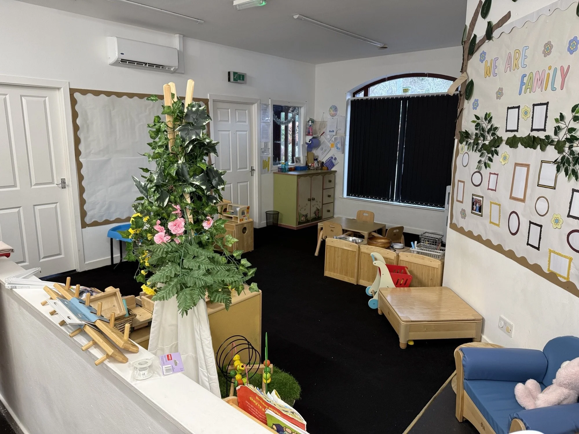 Interior of a preschool classroom with toys, small furniture, and colorful decorations, including a large flower arrangement and a wall display with photo frames and a slogan that reads 'We Are Family'.