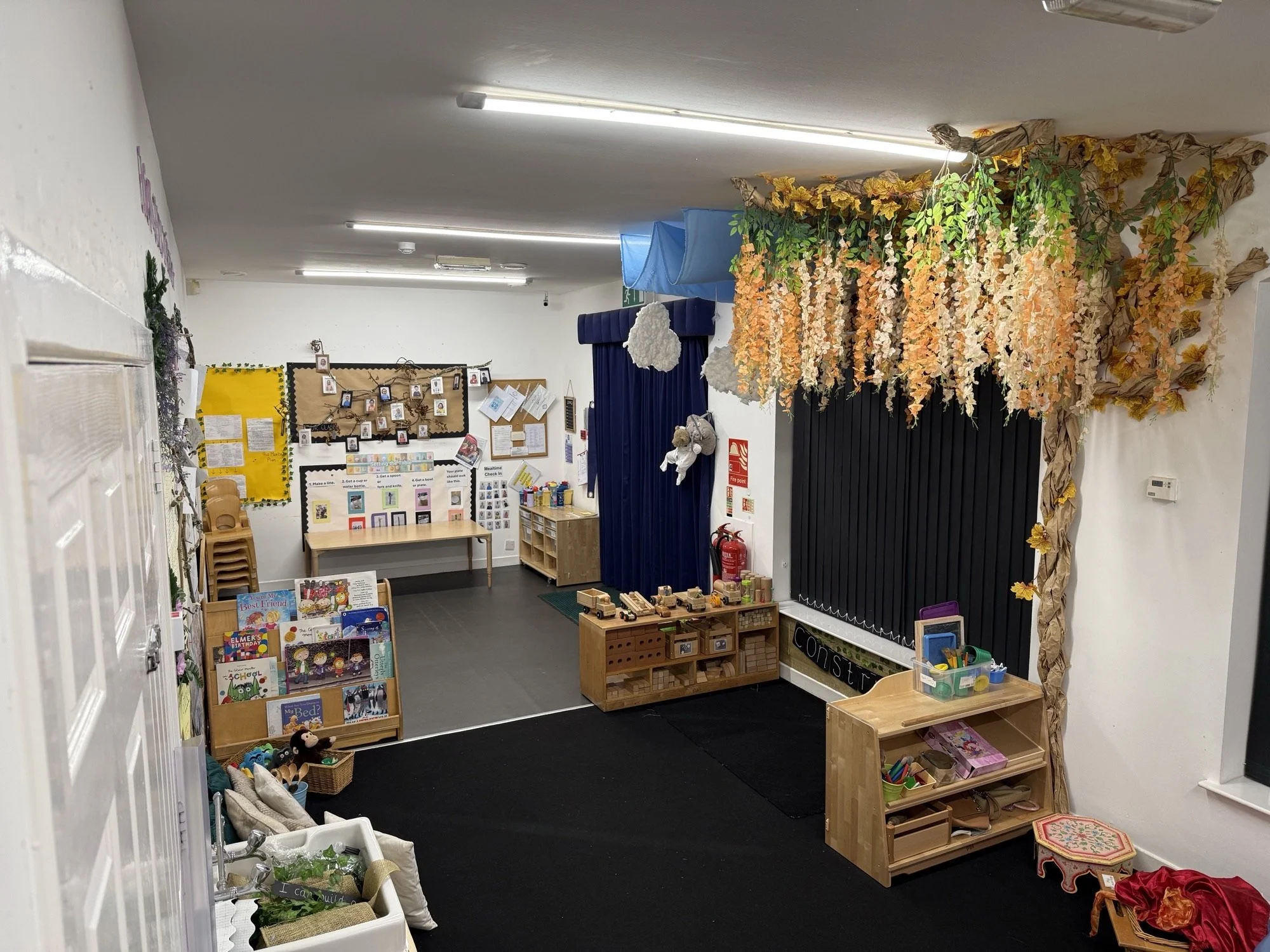 A classroom or playroom with educational and craft materials, books, and decorations including hanging paper flowers and leaves, bulletin boards, and storage shelves.