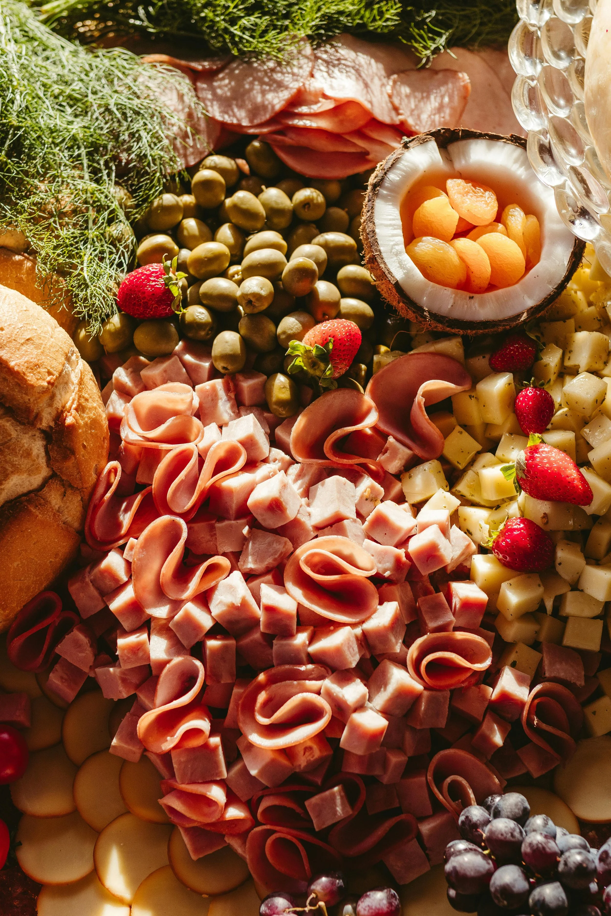 Assorted cheeses, sliced meats, strawberries, green grapes, black grapes, a coconut half with dried apricots, bread, and fresh herbs on a platter.