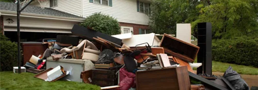Large pile of discarded furniture, including chairs, tables, and sofas, placed in front of a residential house on a lawn, likely for trash pickup.