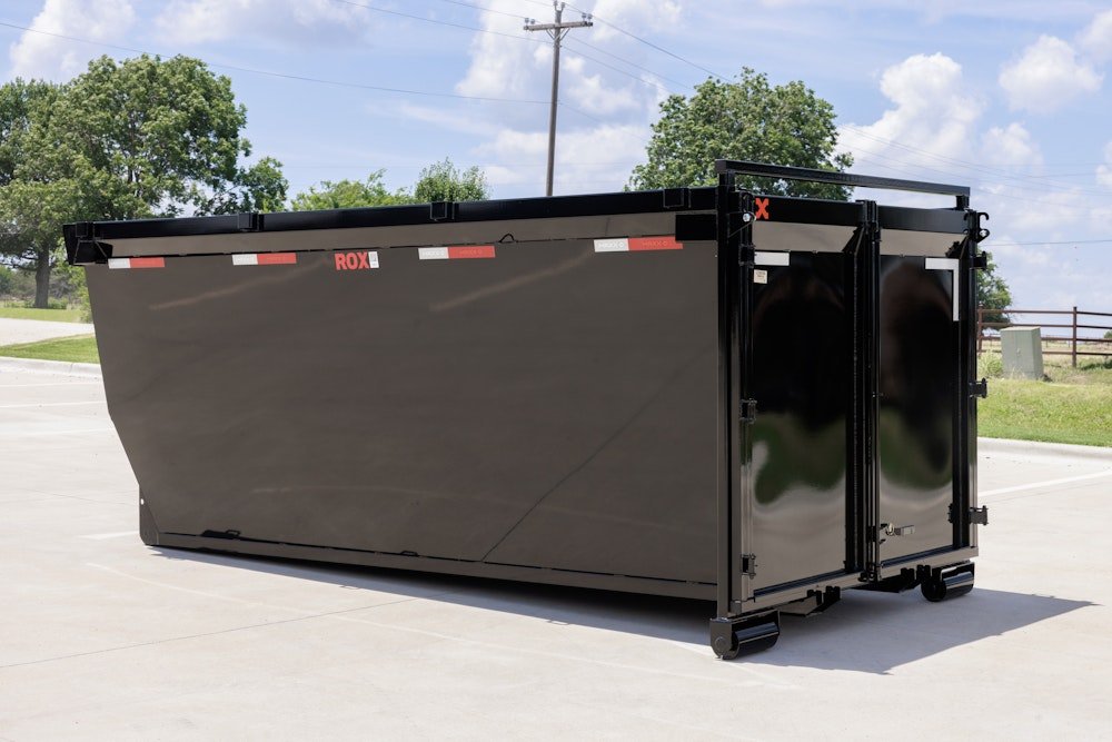 Large black dumpster with red and white reflective strips, placed outdoors on a paved surface, with trees and a blue sky with clouds in the background.