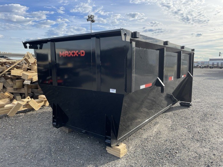 Black MAXX-D dump trailer parked on gravel ground at outdoor storage lot, with wooden blocks supporting it, and a wood pile to the left.
