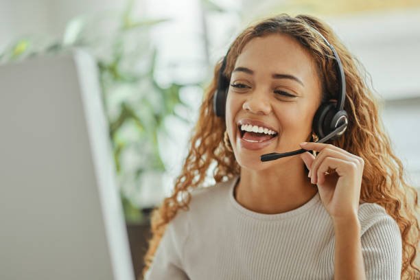 A woman with curly hair wearing a headset, smiling while working on a computer.