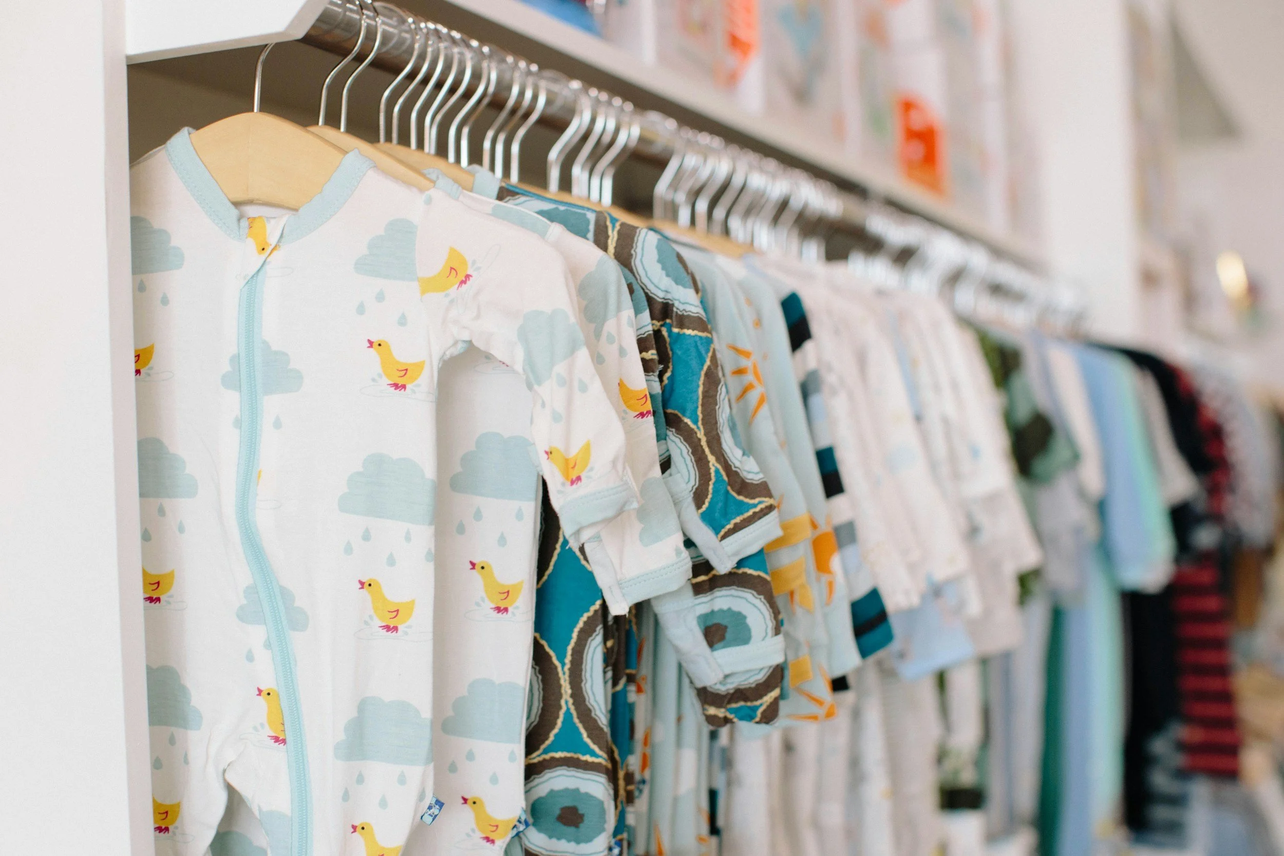 Babies' clothing hanging on a rack, including a white onesie with yellow ducks and gray clouds, and other colorful baby clothes.