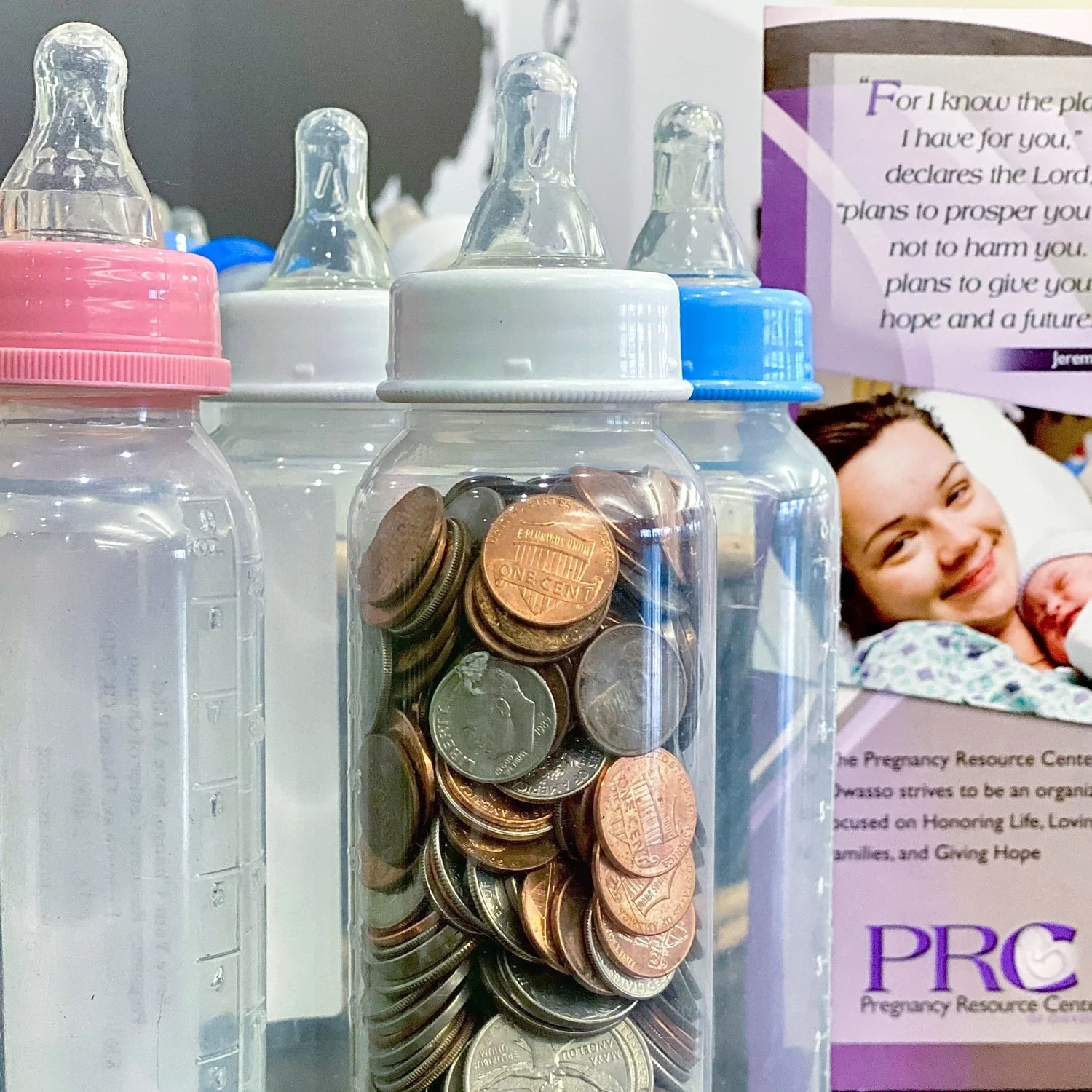 A clear plastic baby bottle filled with coins, with a pink lid and nipple, positioned among other baby bottles on a surface. In the background, a poster with a smiling woman holding a baby is visible, along with a quote and information about a pregnancy resource center.