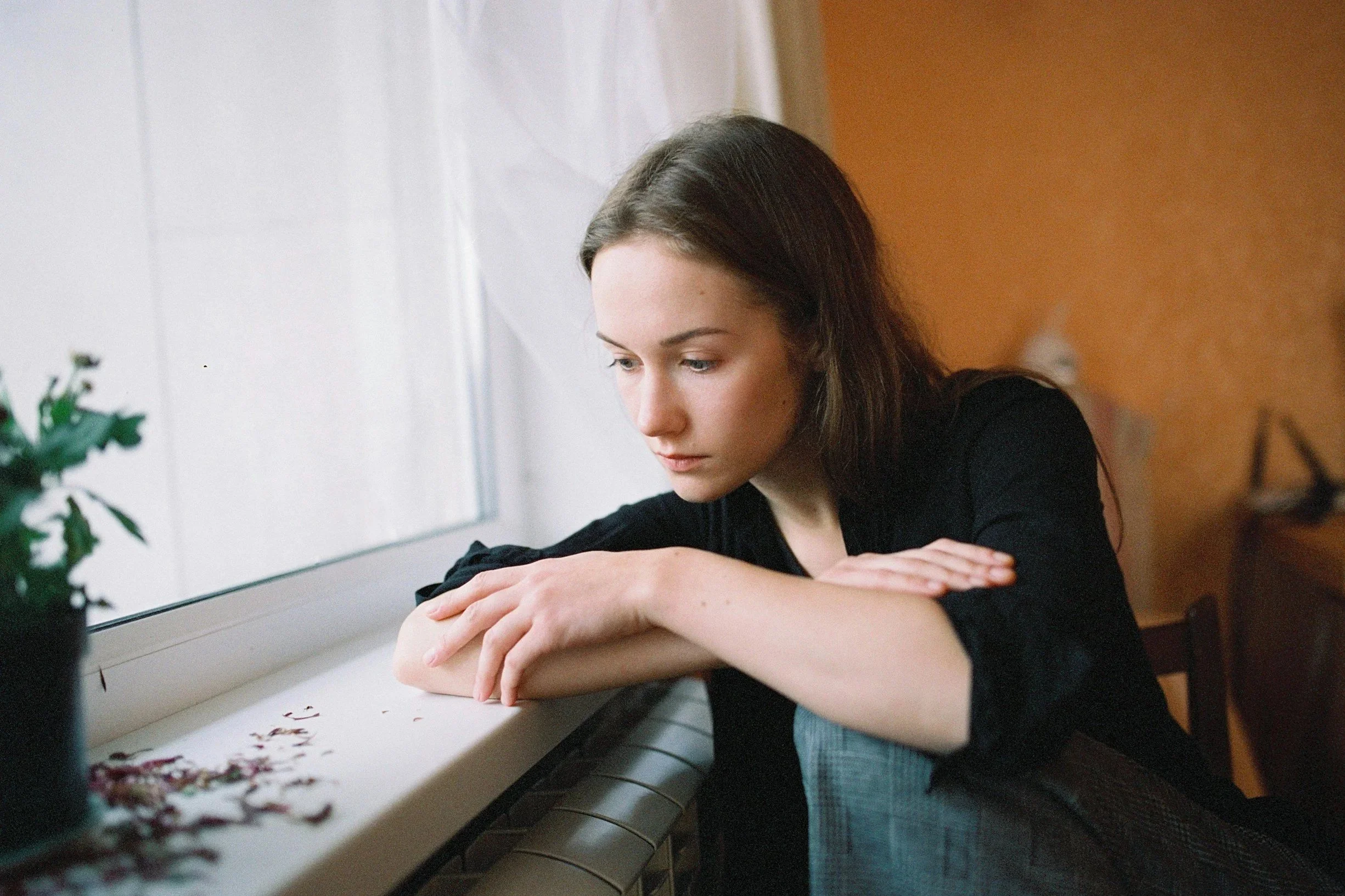 A young woman with brown hair sits by a window, resting her head and arms on the windowsill, looking down with a pensive expression.