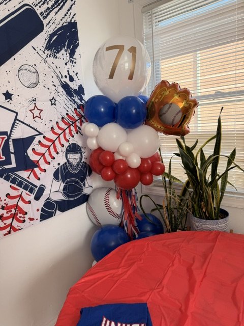 A baseball-themed birthday balloon arrangement with multicolored balloons, including a large clear balloon with the number 71, and a baseball-shaped balloon.
