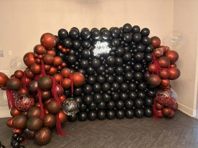 Balloon wall setup with black and red balloons, a neon 'Happy Birthday' sign, and hanging red tassels.