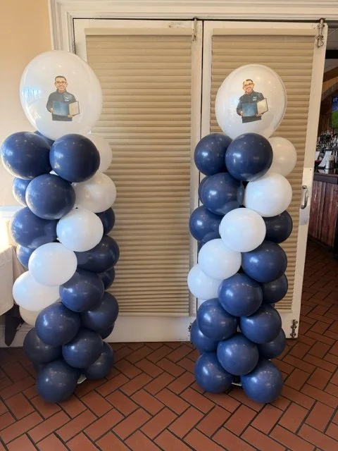 Two balloon columns made of blue and white balloons, topped with round transparent balloons featuring a printed image of a person holding a boxed item, positioned in front of closed louvered doors. Ask us about adding images to balloons!