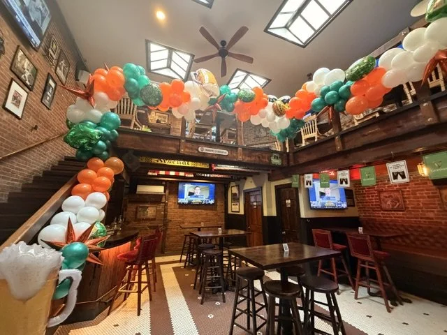 Decorated interior of a restaurant or bar with a large balloon garland arch in red, white, green, and orange, hanging from the ceiling, with tables and chairs, brick walls, and television screens. St. Patrick's day