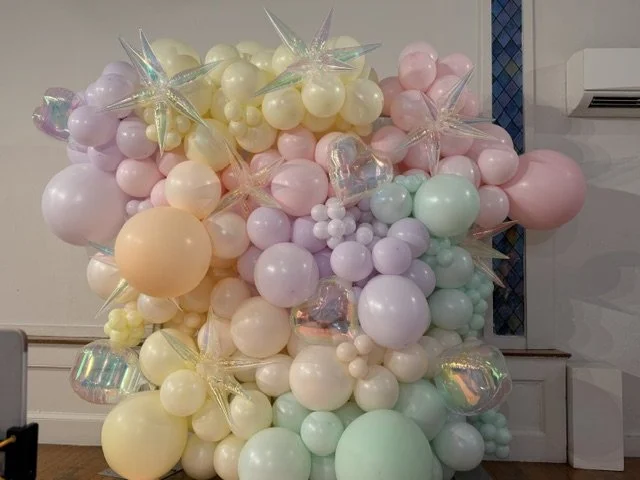 Colorful bunch of pastel balloons in shades of pink, yellow, purple, mint green, and peach arranged in a decorative display against a white wall.
