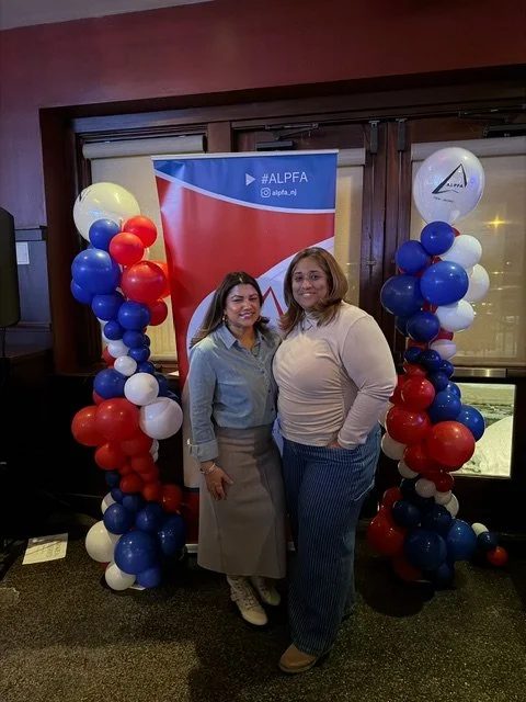 Two women standing between balloon columns with red, white, and blue balloons, in front of a red and blue banner with the hashtag #ALPFA, at an indoor event. We also provide corporate services.