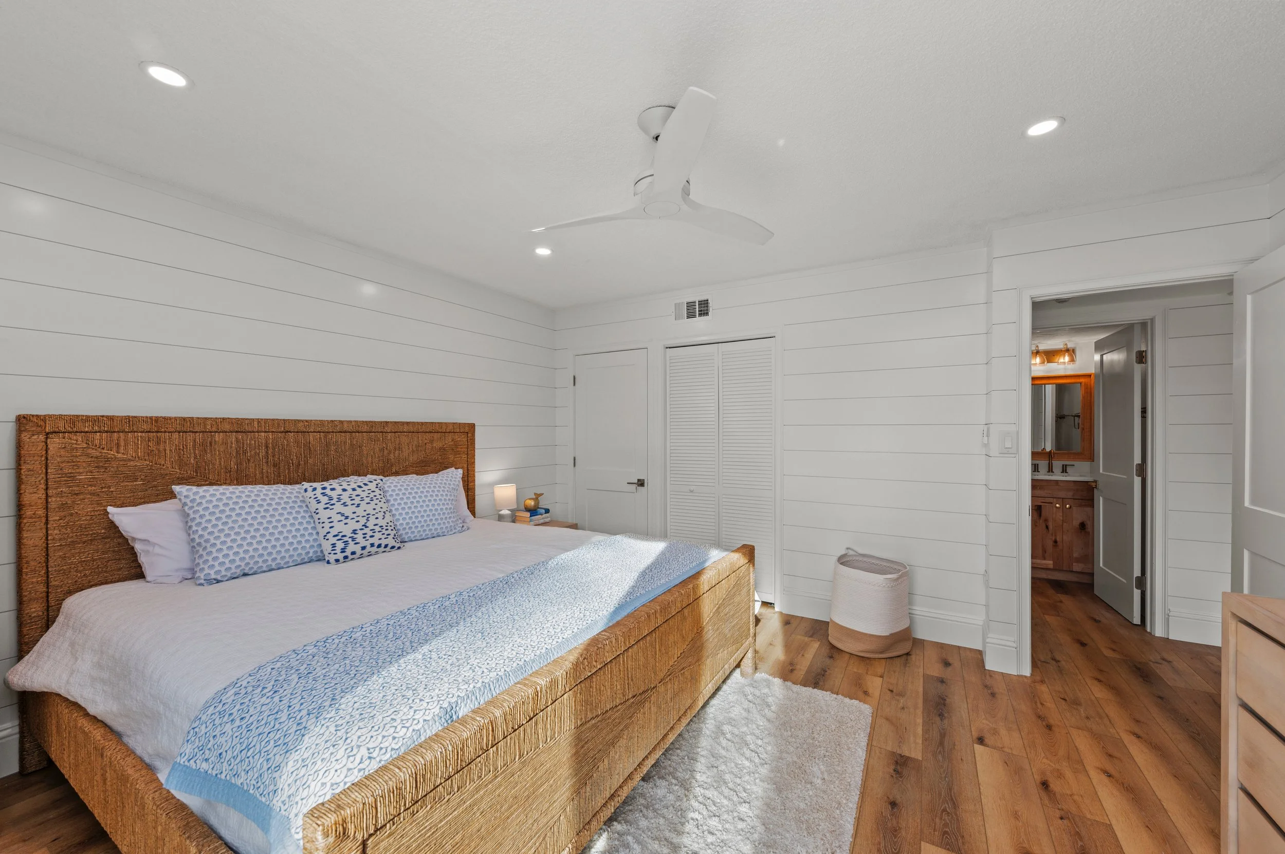 A bedroom with a king-sized bed, wooden headboard, white bedding, and blue pillows, sunlight on the floor, white walls, ceiling fan, door to a bathroom with wooden vanity and mirror, woven laundry basket, and a small side table with a lamp.