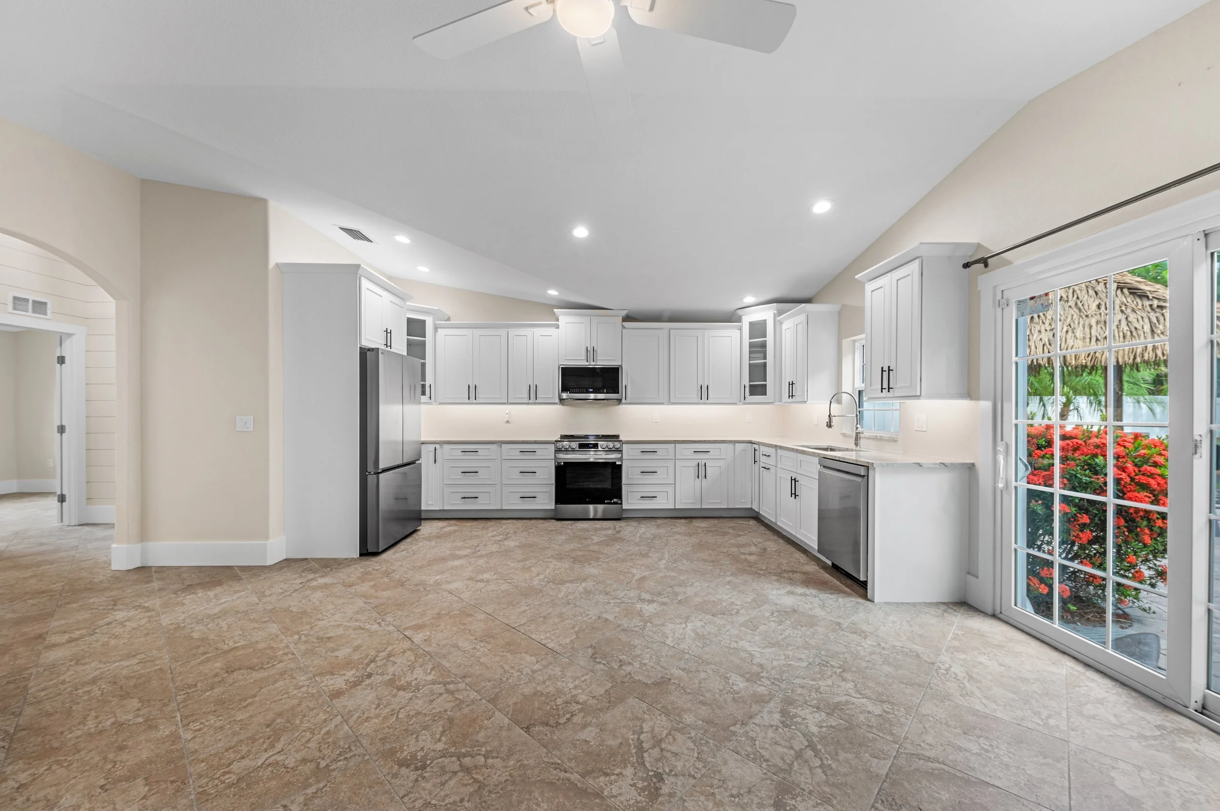 Modern kitchen with white cabinets, stainless steel refrigerator, oven, dishwasher, and a large sliding glass door leading outside to a garden with red flowers and a thatched roof.