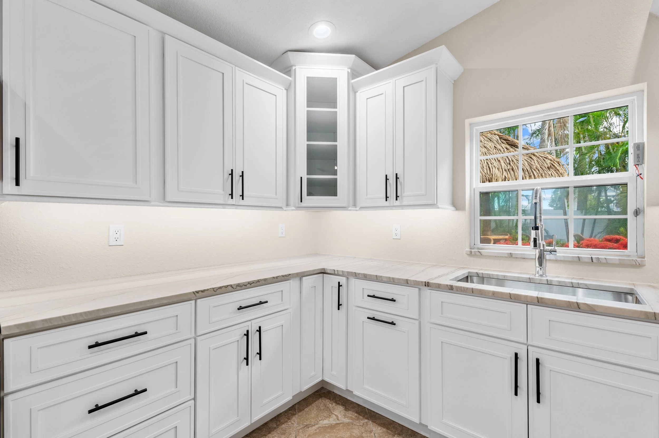 Modern white kitchen with upper and lower cabinets, a marble countertop, and a window overlooking a tropical outdoor scene.