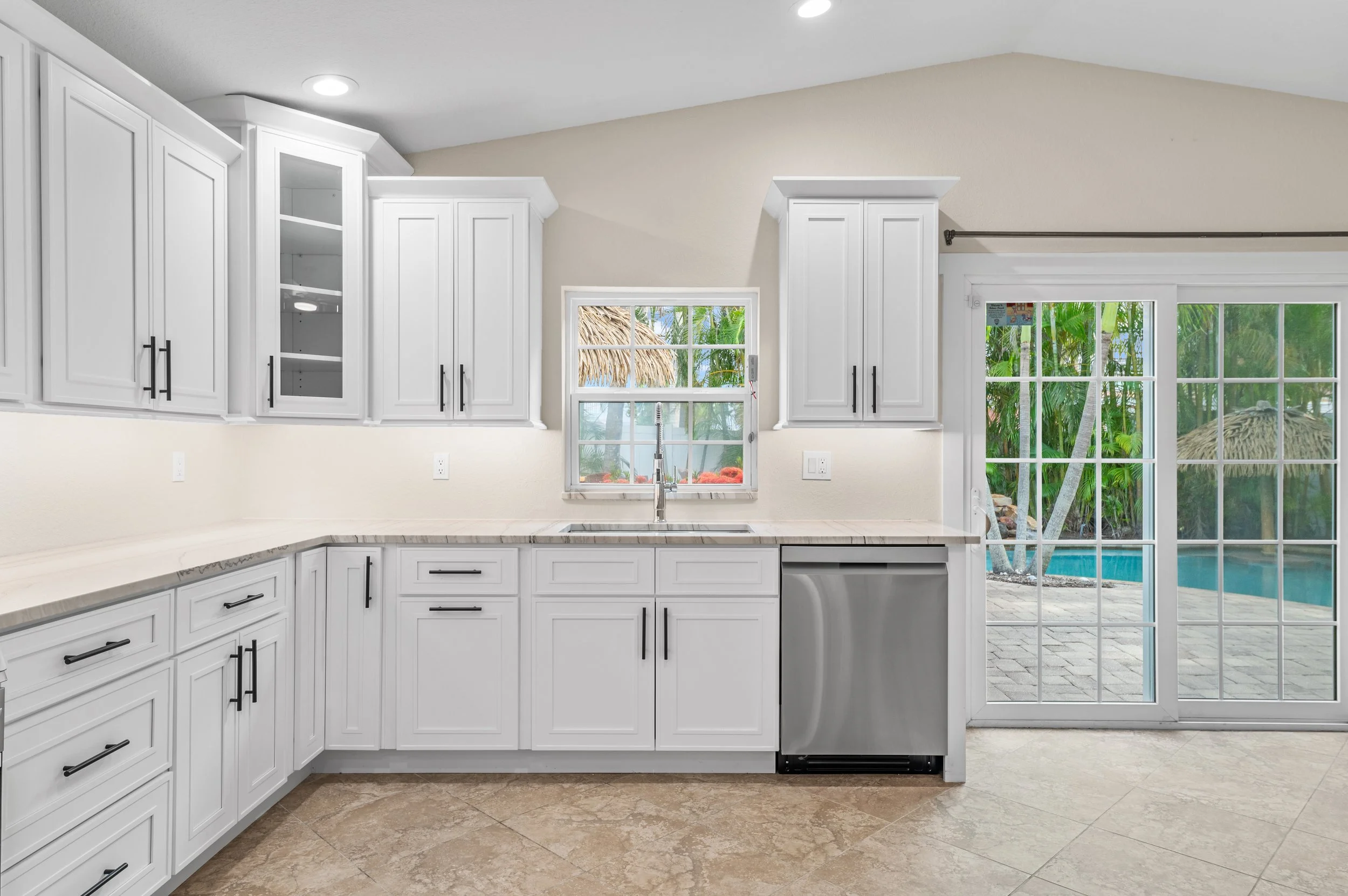 Kitchen with white cabinets, marble countertop, window above the sink, sliding glass door to outside patio, tropical trees and pool visible through the door.