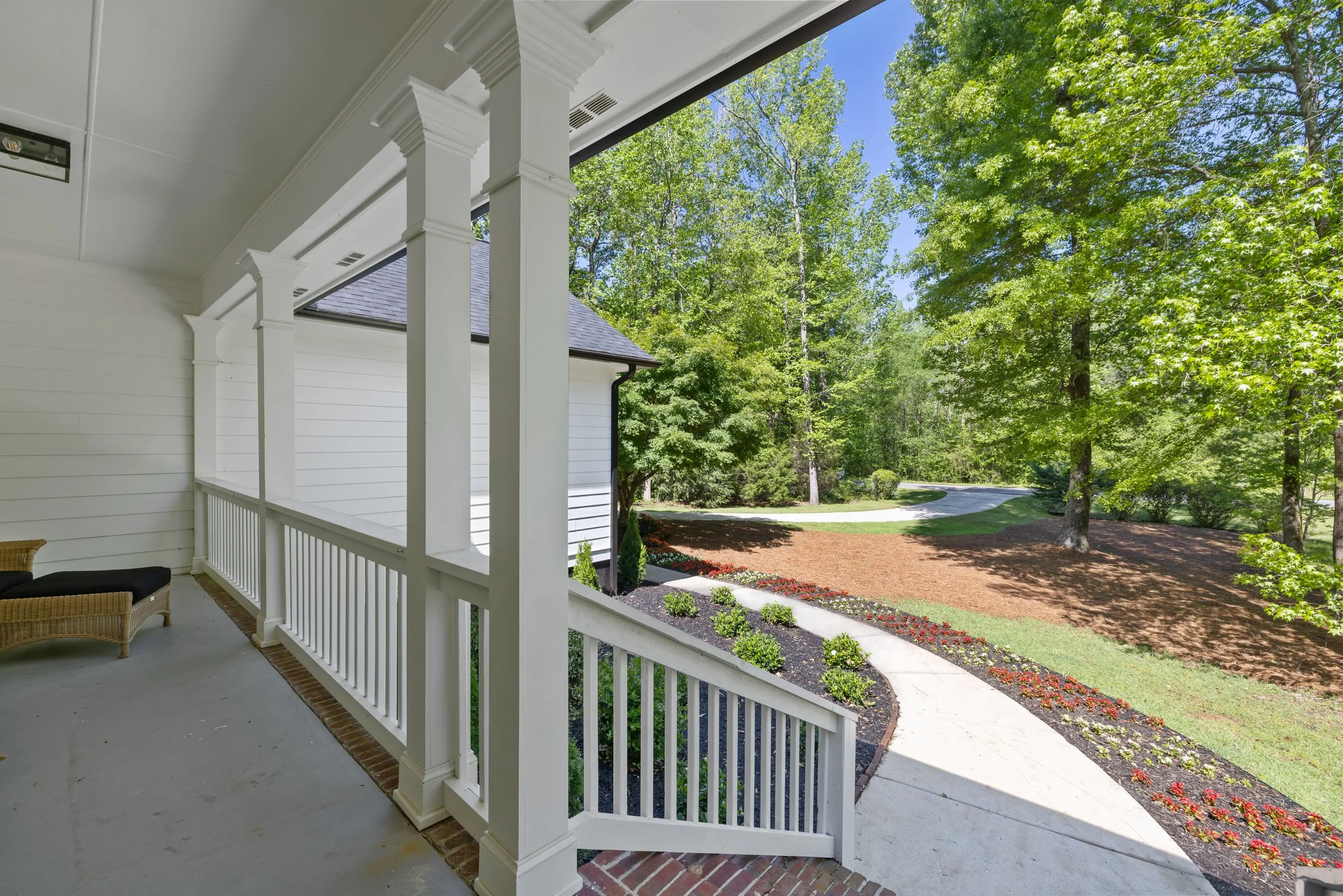 View from a porch showing a curved walkway, landscaped garden beds with flowers, and green trees under a bright blue sky.