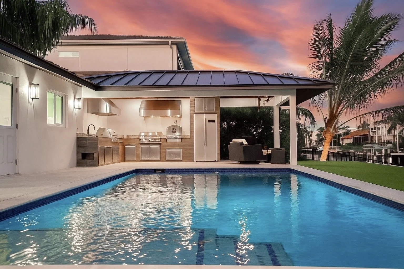 Modern backyard with a swimming pool, outdoor kitchen, and seating area under a covered patio, with a sunset sky and palm trees in the background.